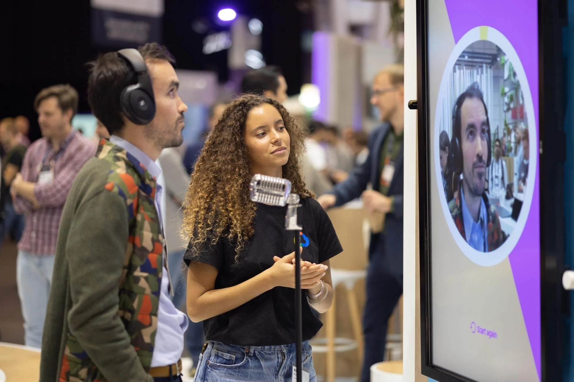 A woman with curly hair is speaking into a microphone while a man wearing headphones listens at a tech conference or exhibition.
