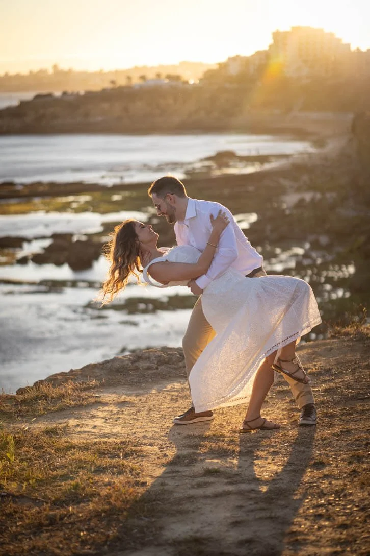 A couple dancing on a rocky beach at sunset, with water and distant buildings in the background.