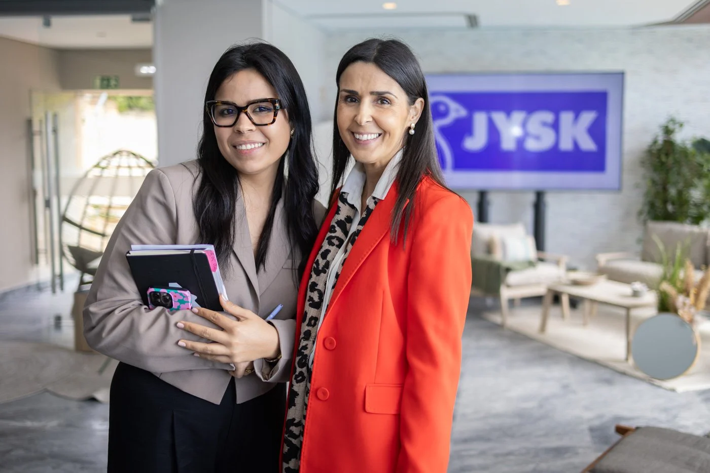 Two women standing in front of a JYSK sign, smiling at the camera, indoors in a furniture store or showroom.
