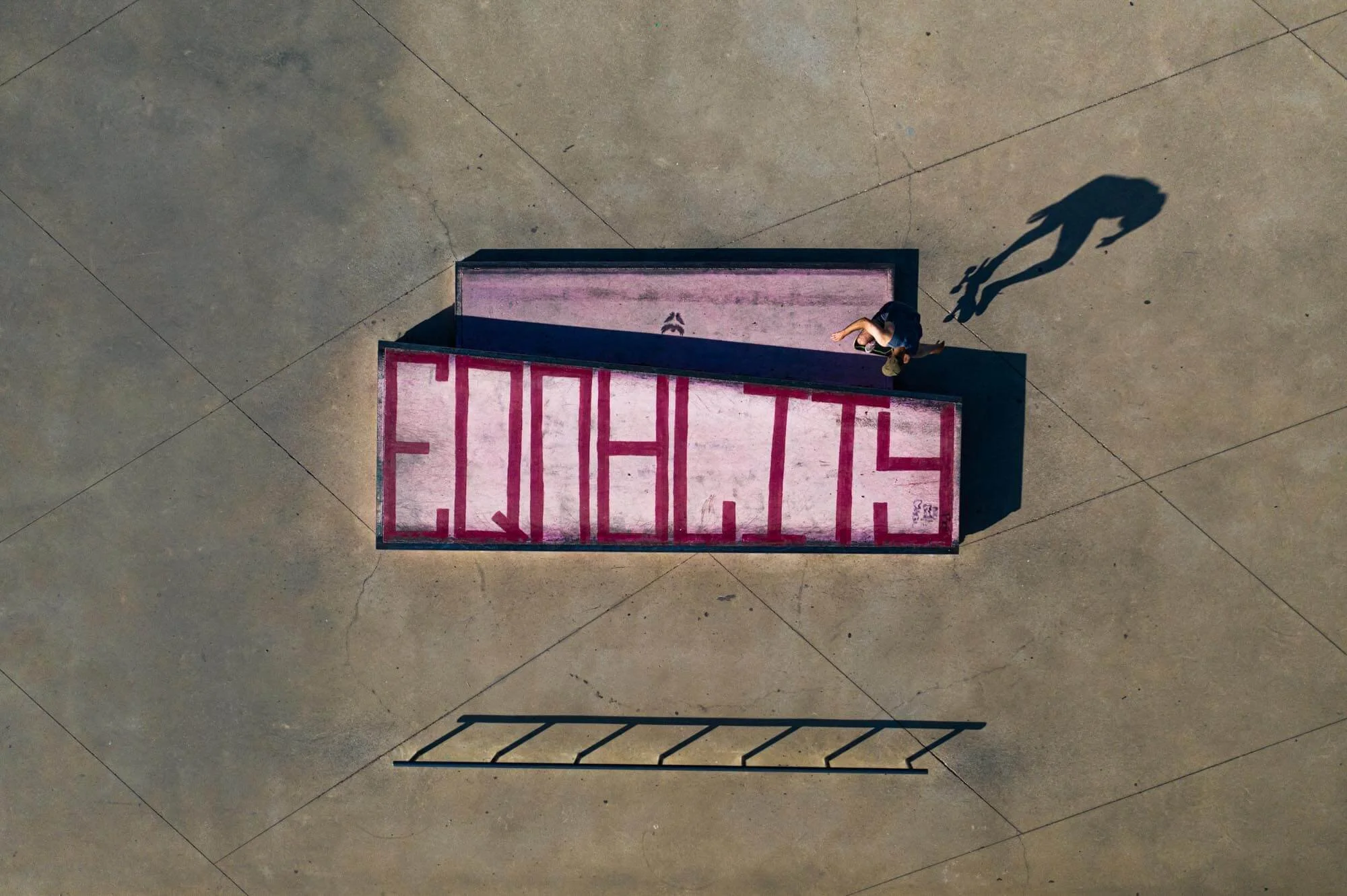 Top-down view of a man looking at a large pink sign with red letters spelling 'ERHGM' on a concrete surface with a shadow of a ladder below.