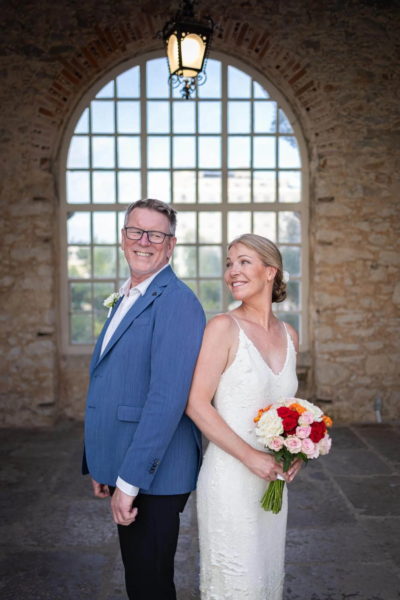 A bride in a white wedding dress holding a bouquet of pink, red, and white roses, standing back-to-back with a groom in a blue suit, smiling, inside a rustic stone building with a large arched window and hanging lantern.