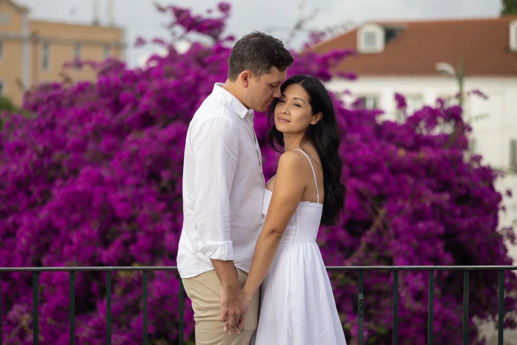 A couple holding hands and leaning close together in front of a large purple flowering bush, with buildings in the background.