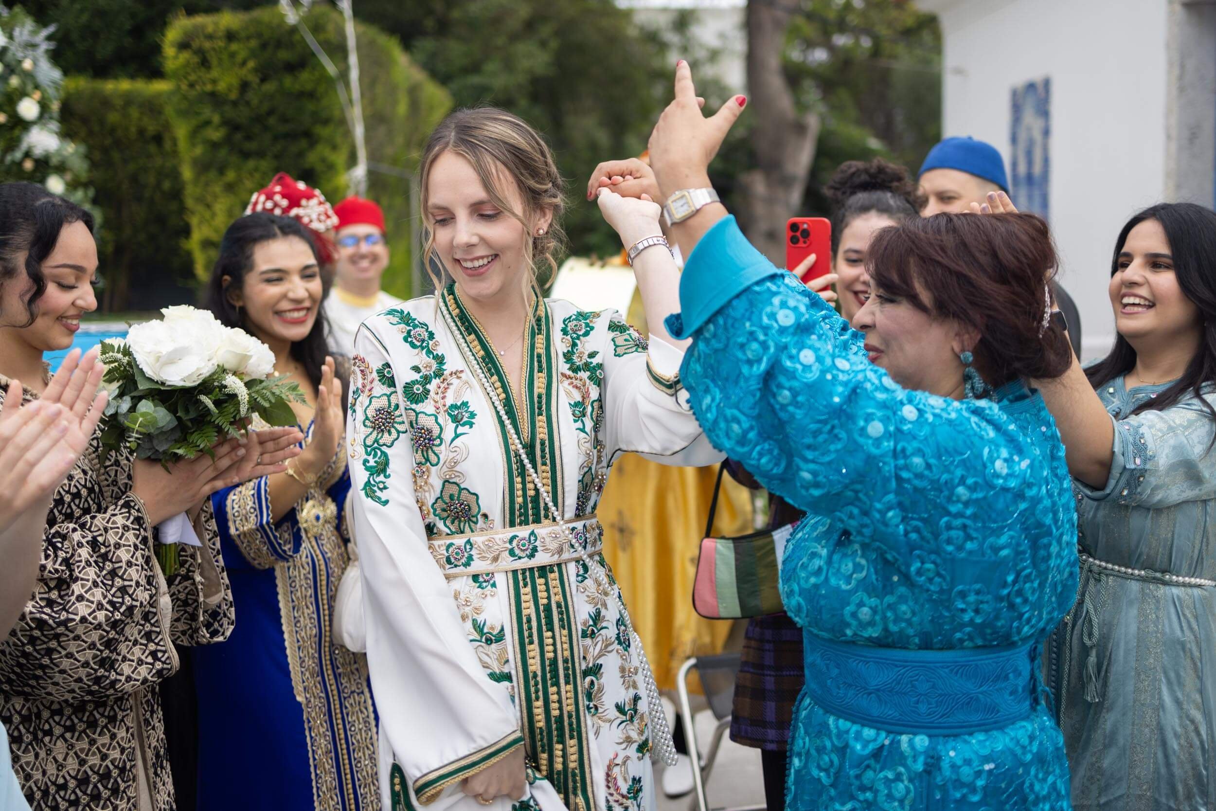 Group of women in traditional clothing celebrating outdoors, with one woman holding a bouquet of white flowers.