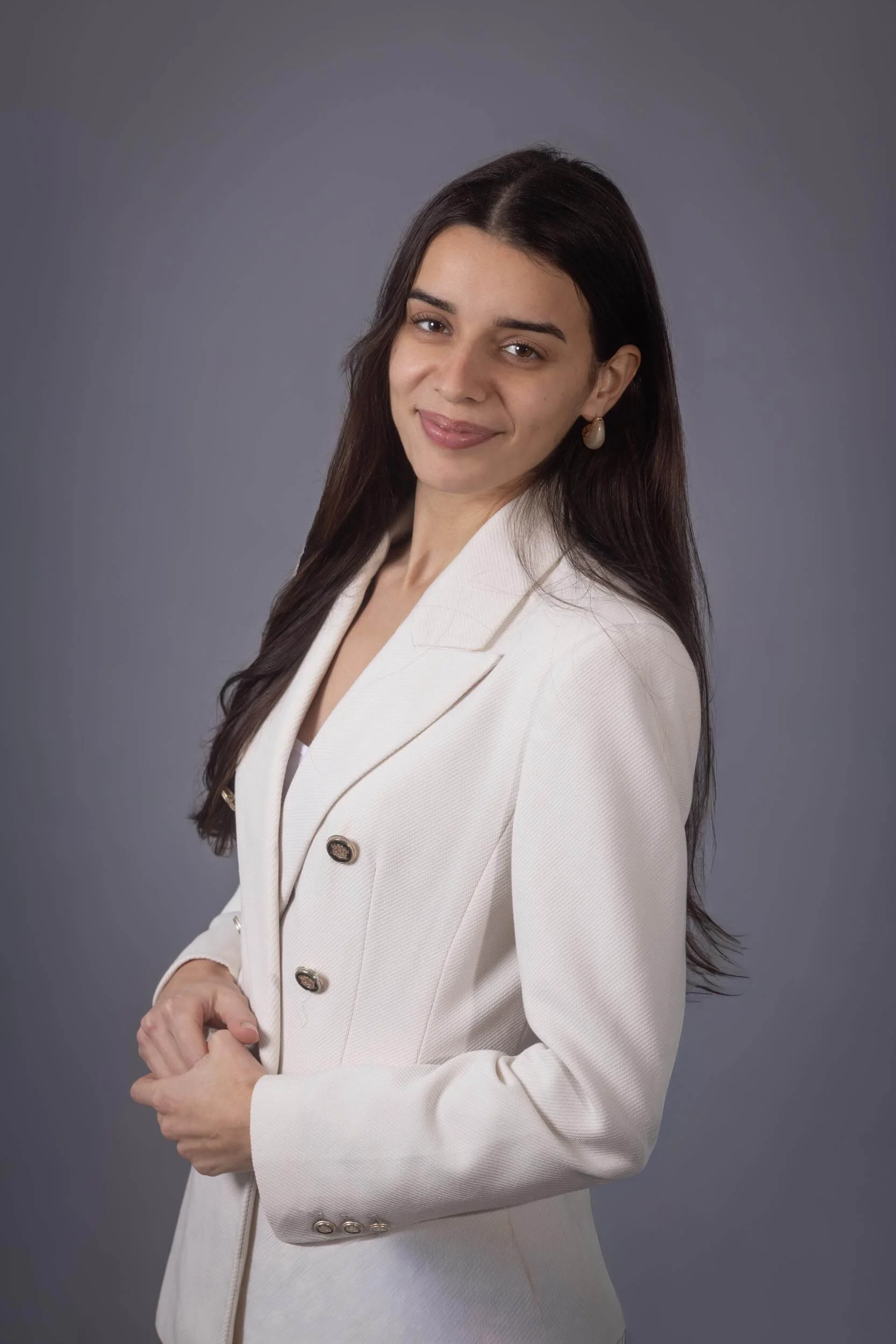 A young woman with long dark hair, wearing a white blazer, smiling and looking at the camera against a gray background.