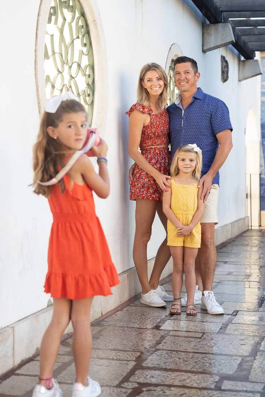 A family of four posing on a sidewalk against a white wall with decorative windows. The mother and father stand close together, with the mother wearing a red floral dress and the father in a blue patterned shirt and shorts. Two young girls, one in a 