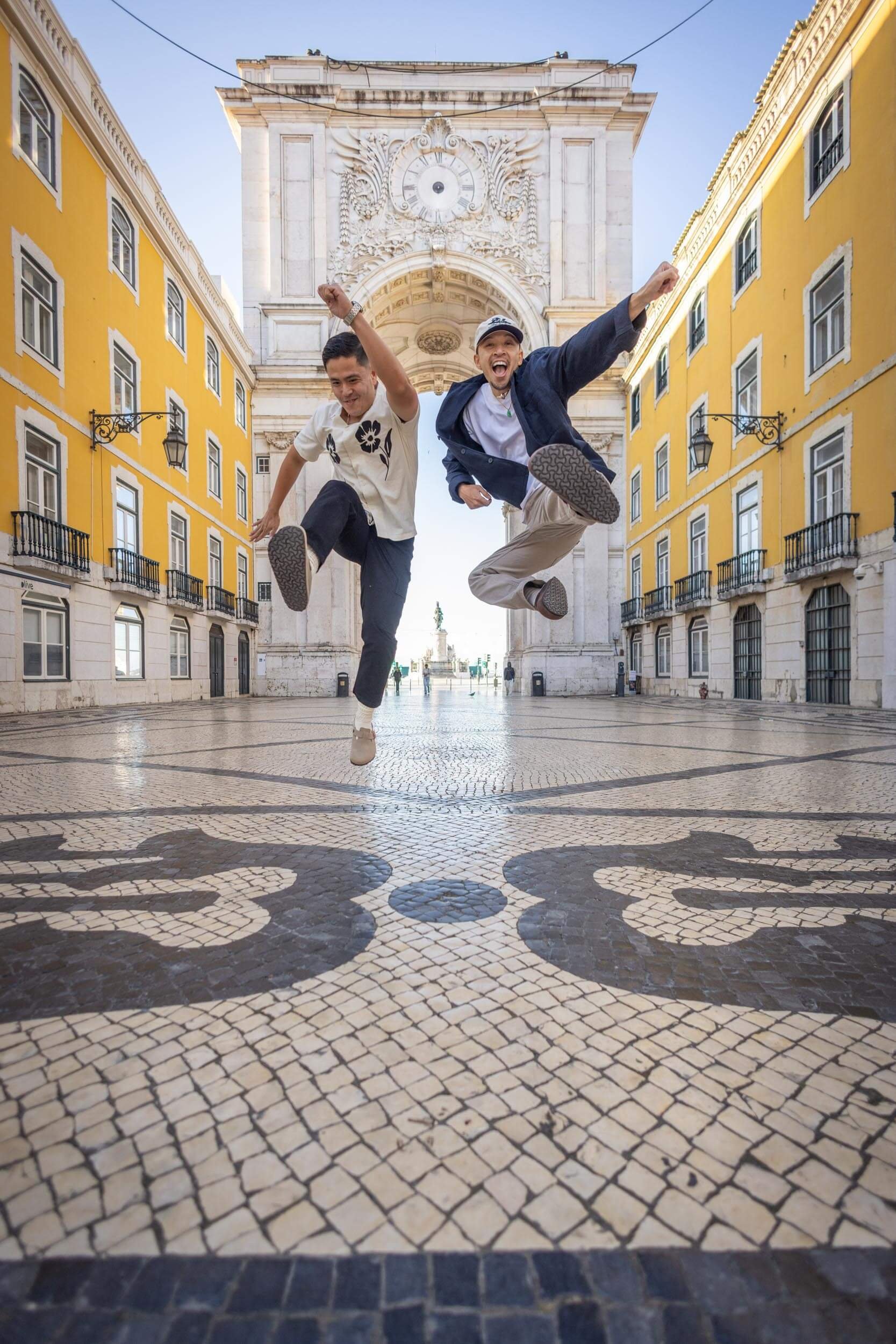 Two young men, a couple,  are jumping in the air and smiling in a  Lisbon historic square with yellow buildings and an ornate archway in the background.