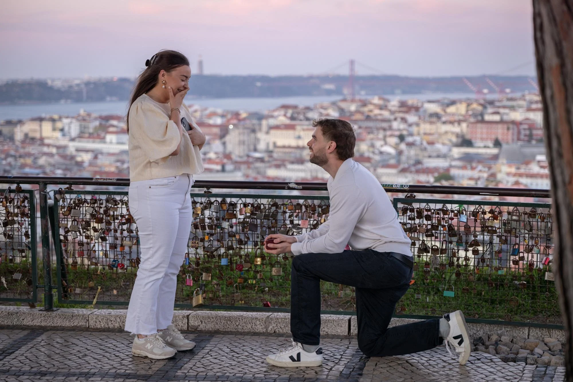 A man proposing to a woman on a balcony overlooking a cityscape and a bridge at sunset.