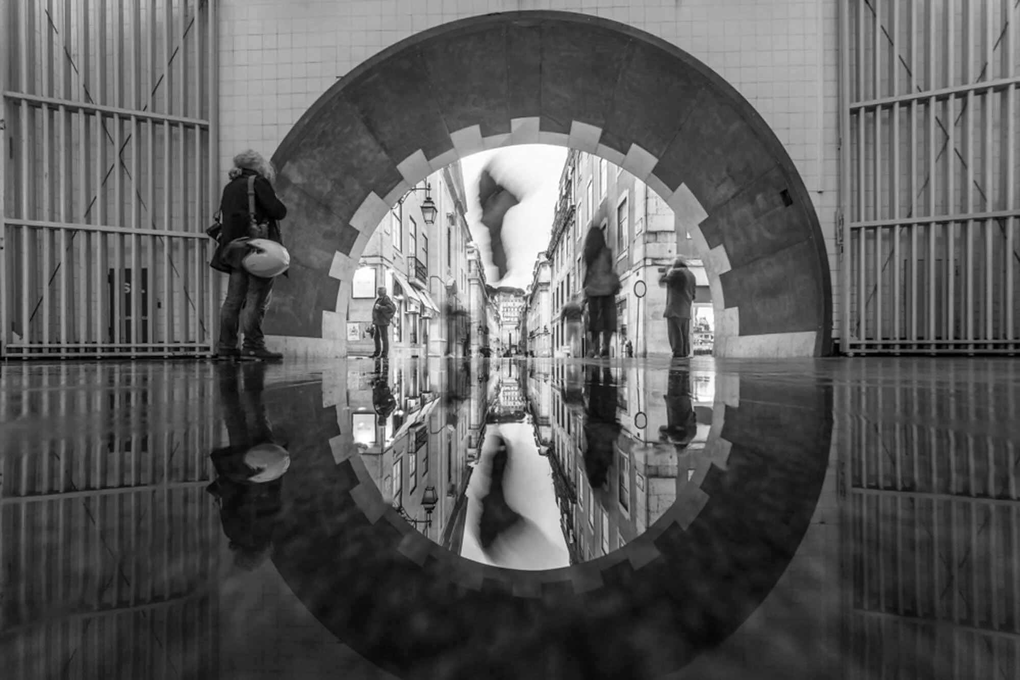 Black and white photo of a city street viewed through a circular mirror, reflecting pedestrians walking and signage.