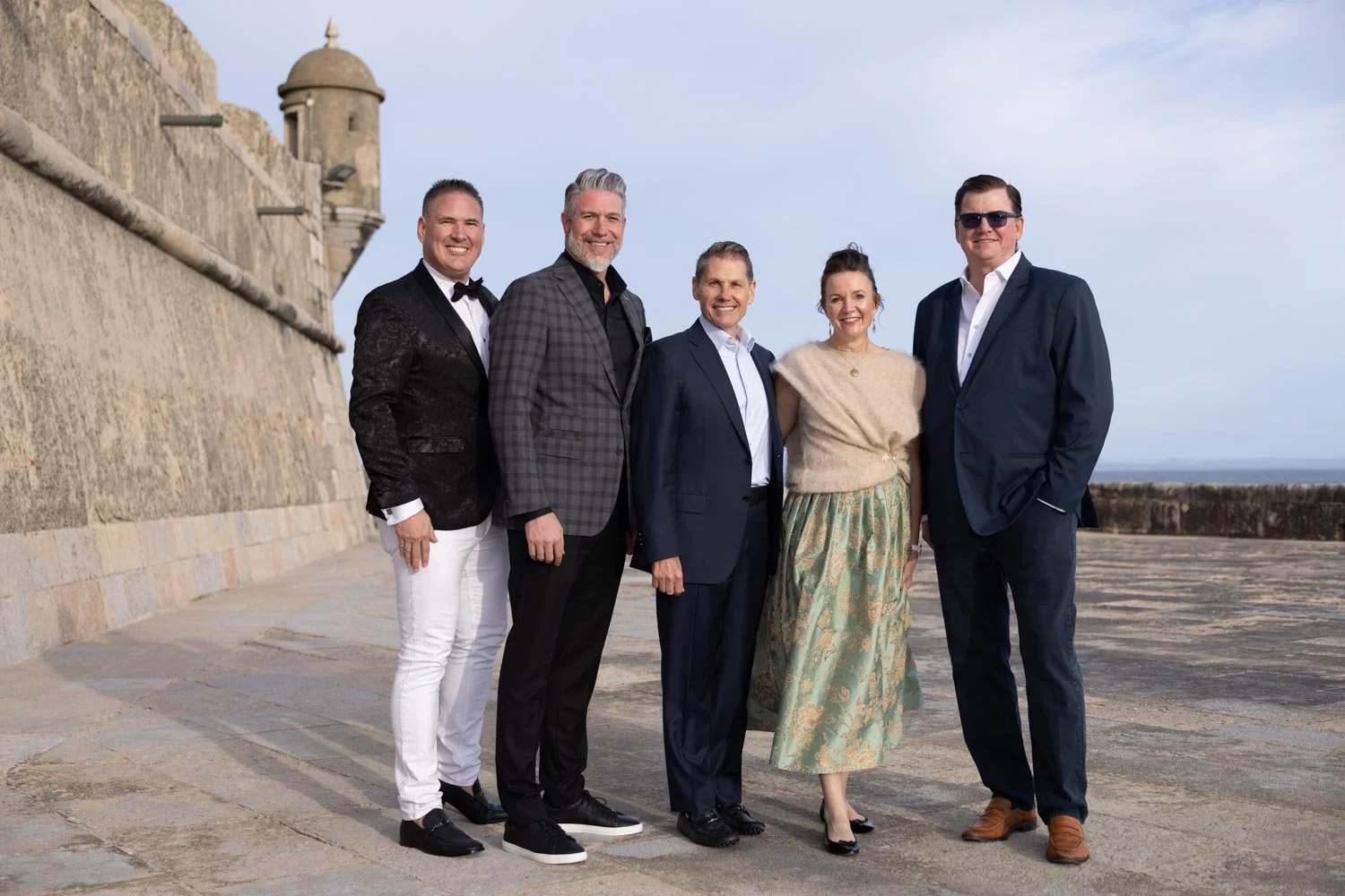 Group of six people dressed in formal and semi-formal attire, standing outdoors near a historic stone wall overlooking the ocean on a clear day. They are posing for a photographer.