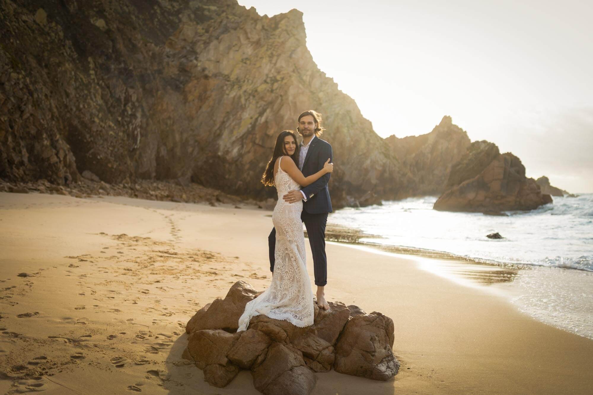 A couple dressed in wedding attire standing on rocks on a sandy beach with cliffs in the background at sunset.
