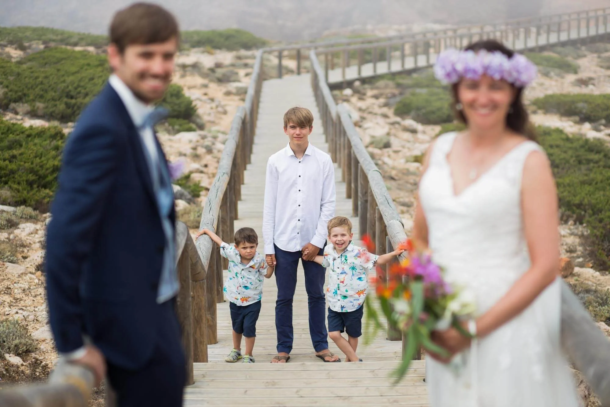 A wedding scene on a wooden bridge with a bride holding a bouquet of flowers in the foreground, and three boys and a young man standing in the background, with the young man holding the youngest boy's hand. The scene is outdoors with a rocky, shrub-c