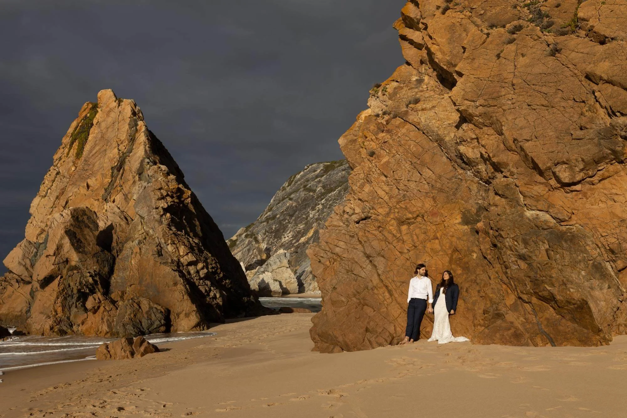 A man and woman holding hands on a sandy beach, standing near large orange-brown rocks with a stormy sky overhead.