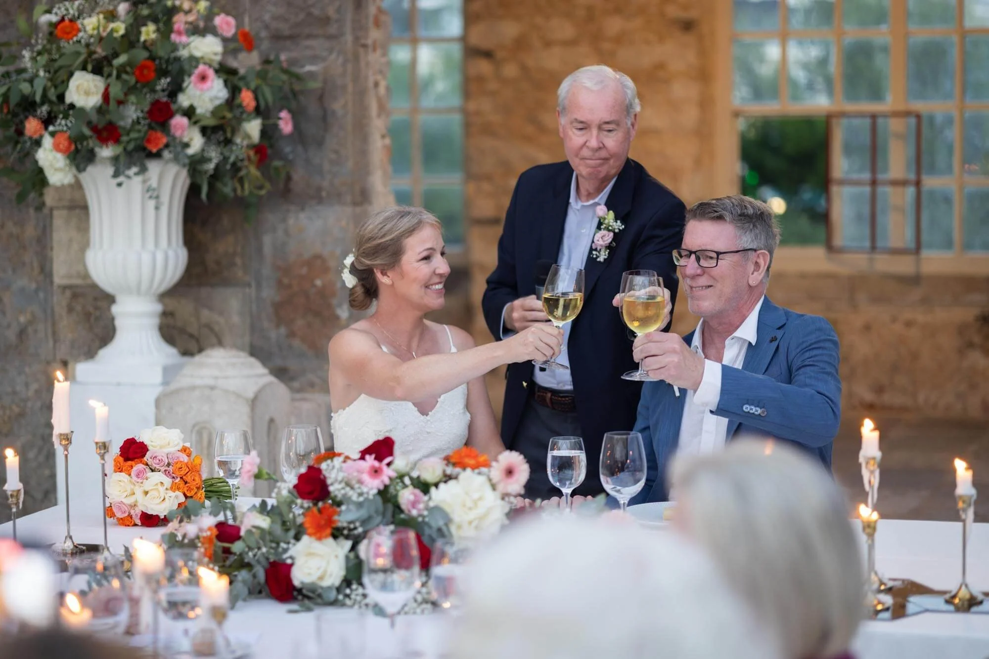 Wedding celebration with three people raising glasses of white wine in a toast. The bride, in a white dress, and the groom, in a blue suit, sitting at a decorated table with flowers and candles, while an older man standing behind them toasts with the