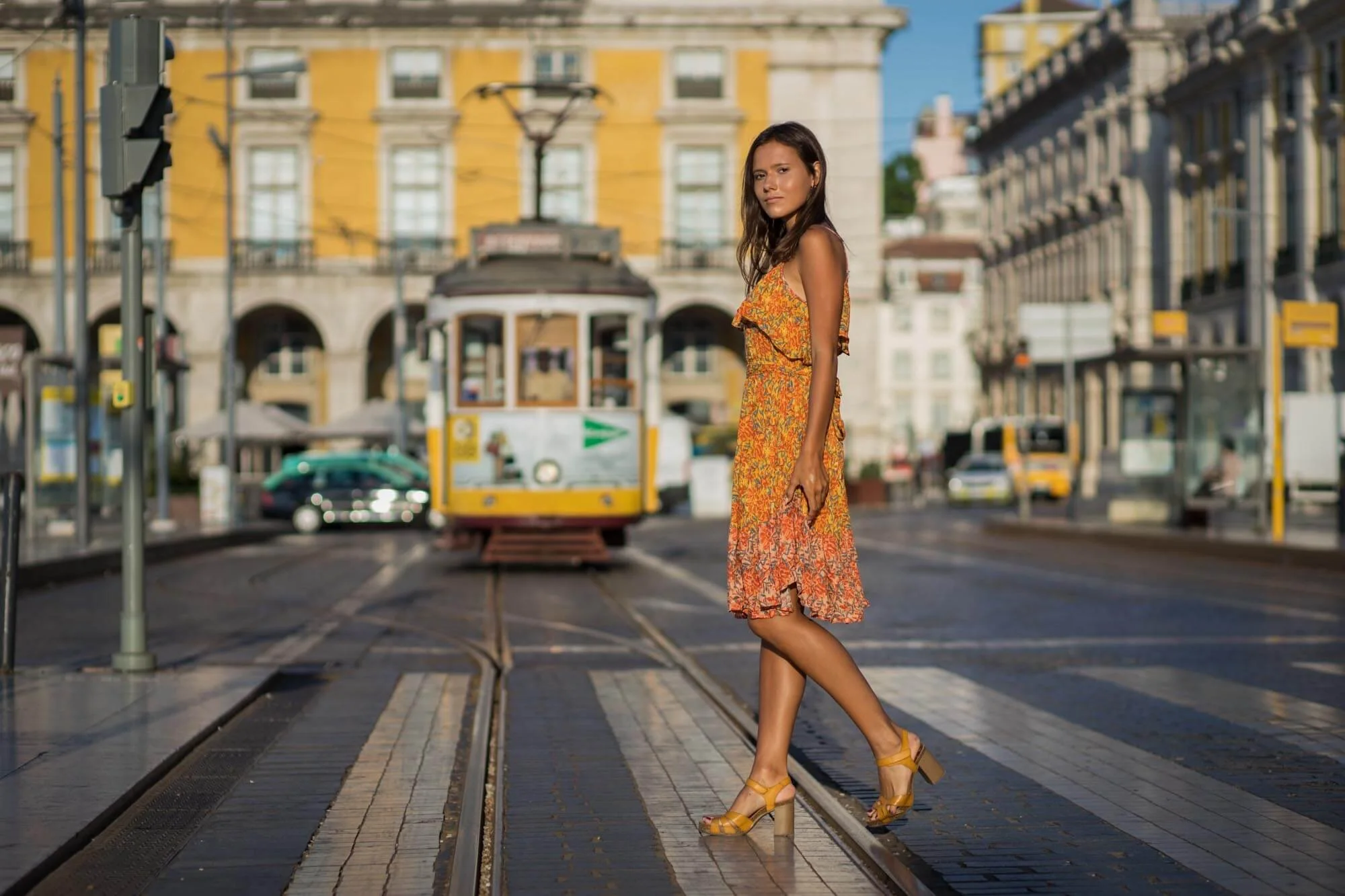A woman in a yellow floral dress and yellow high-heeled sandals standing on tram tracks in an urban setting with tram in the background.