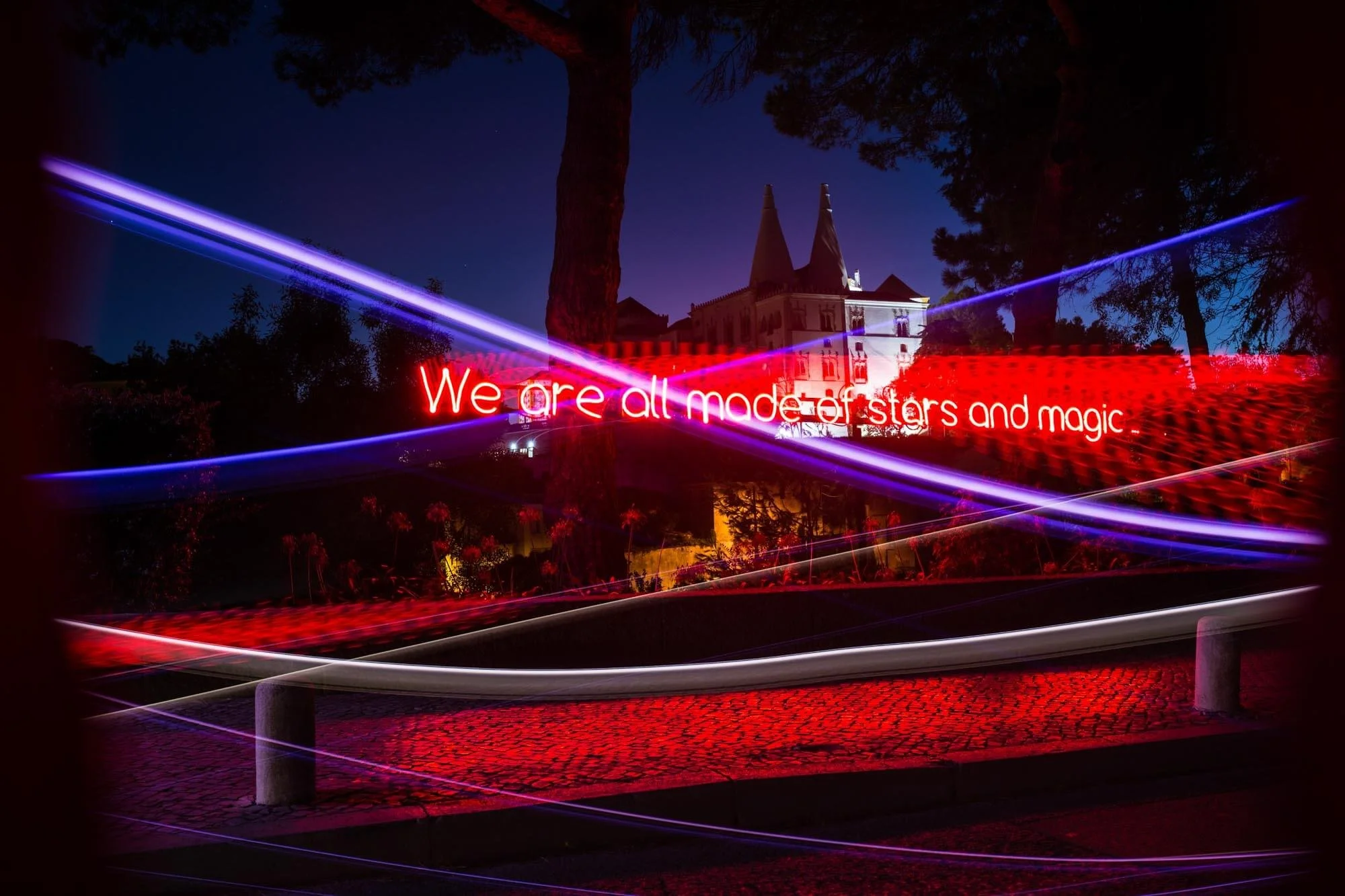 Nighttime scene showing a building with twin towers, trees, and colorful light trails with a red illuminated message: 'We are all made of stars and magic'.