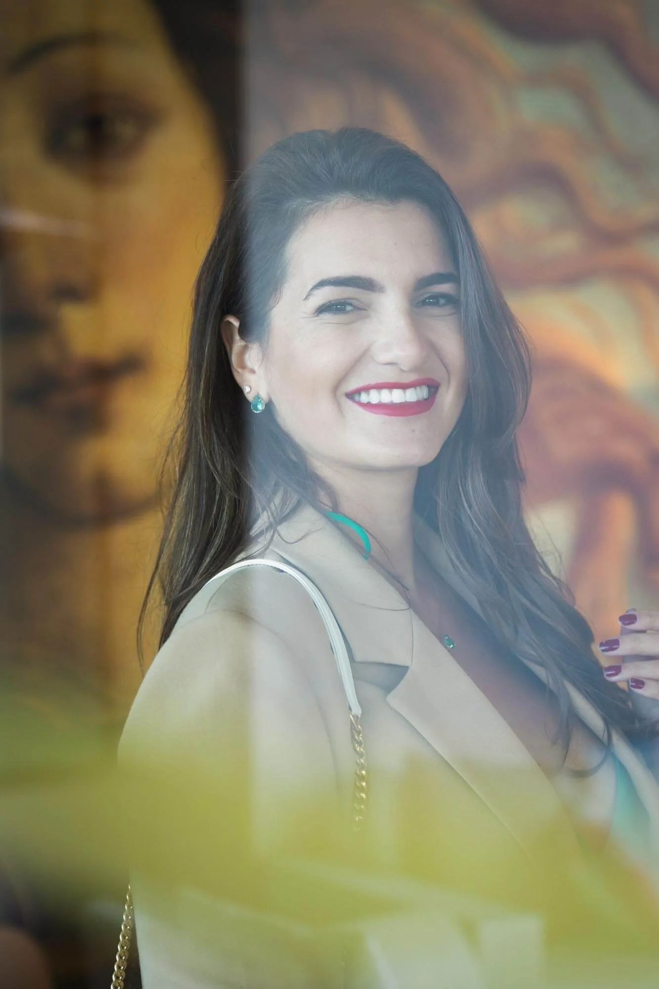 Photo of a smiling woman with dark hair and red lipstick, wearing earrings and a beige blazer, seen through a glass window with a reflection of artwork.