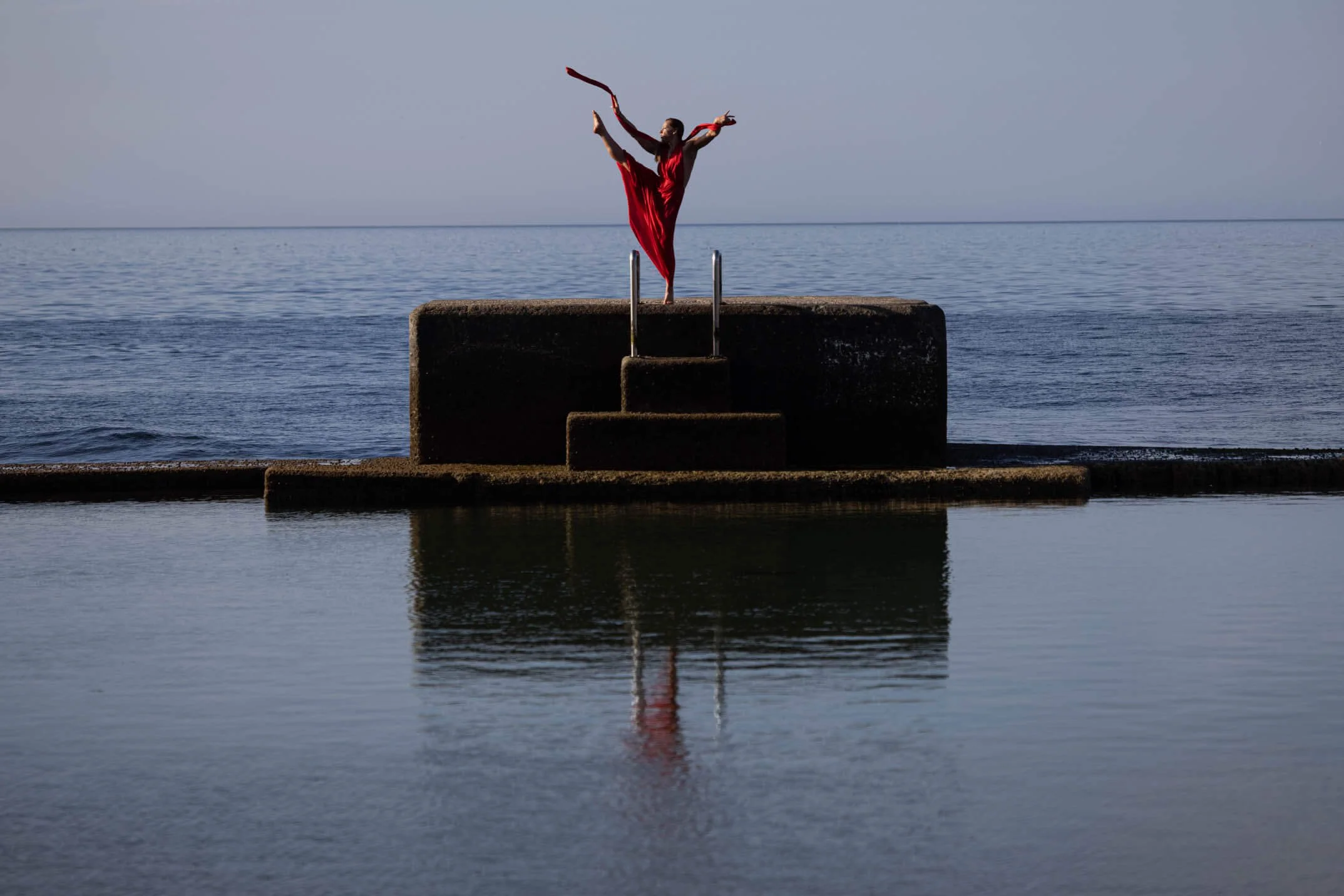 Person in a red dress performing a dance jump on a concrete platform at the edge of the water, with a calm body of water and a cloudy sky in the background.