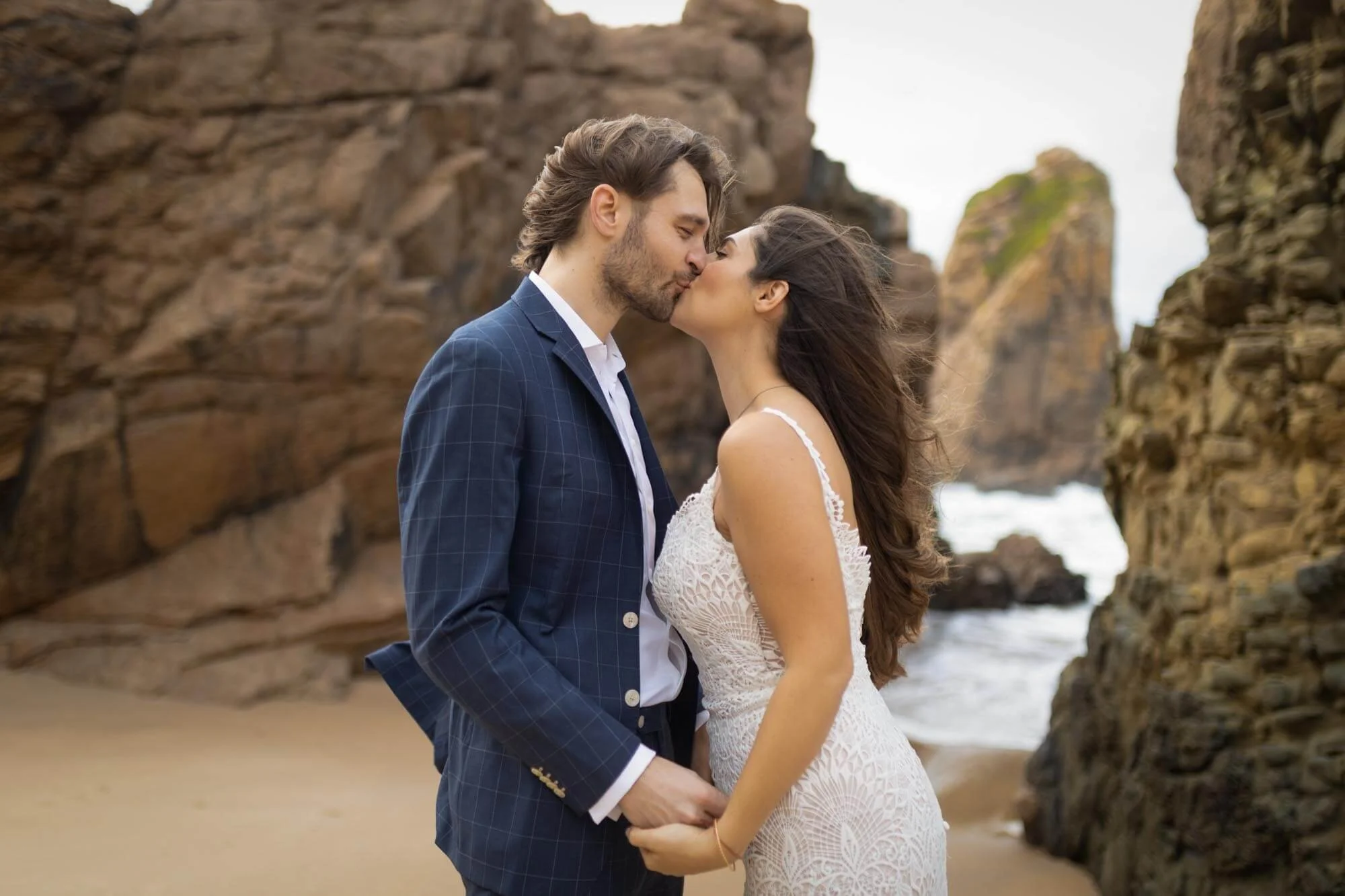 A couple kissing on a beach with rocky cliffs in the background.