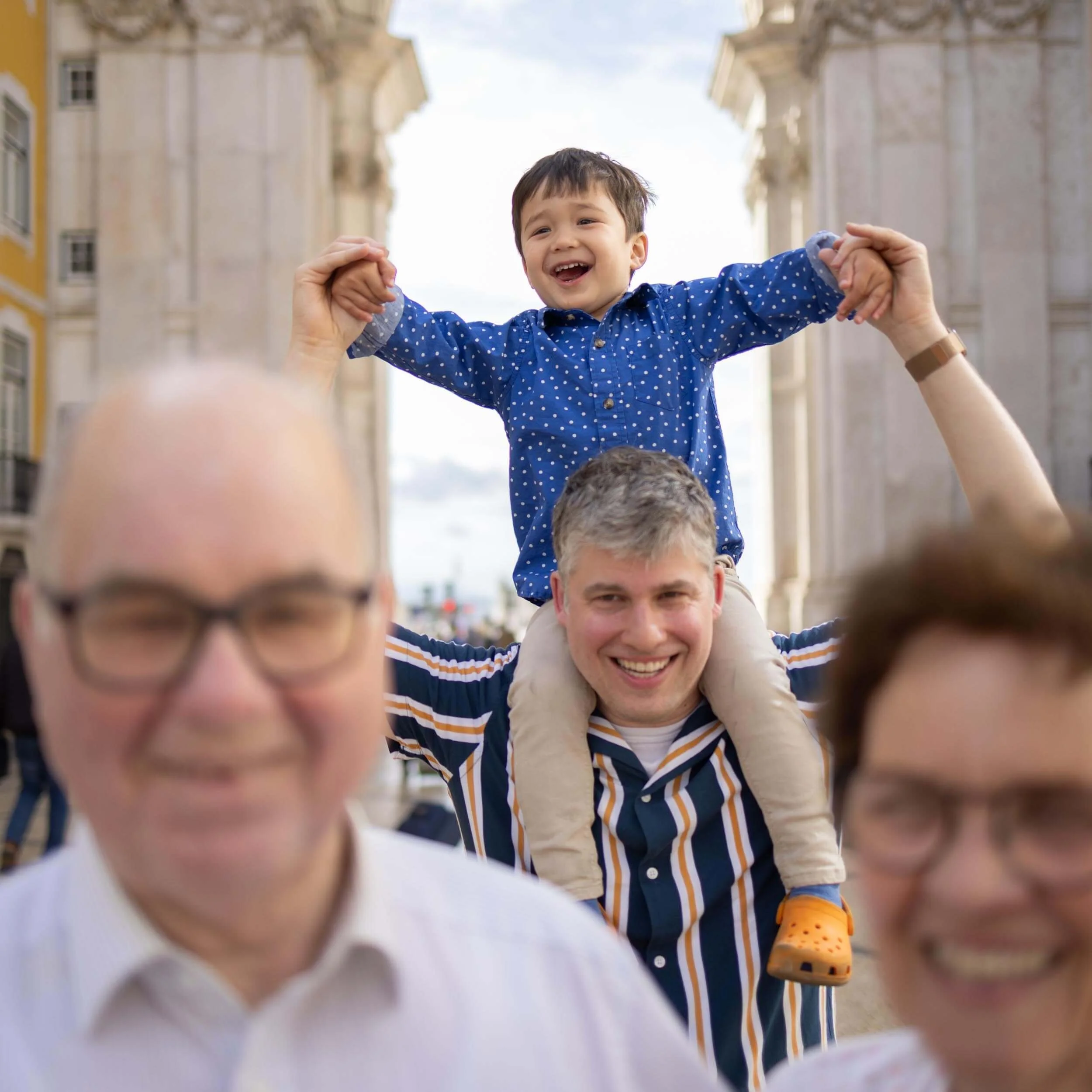 Family enjoying outdoor city sightseeing, with a young boy on adult's shoulders raising his arms in joy.