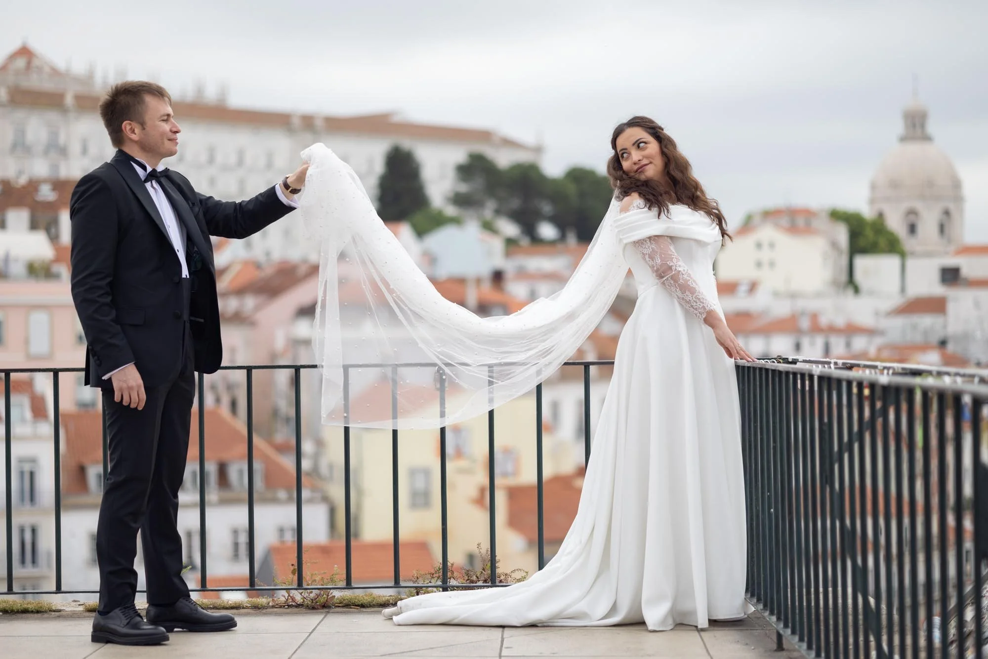 A bride in a white wedding dress with lace sleeves holding onto a railing, and a groom in a black tuxedo holding a sheer veil, standing on a balcony with a cityscape and dome in the background.