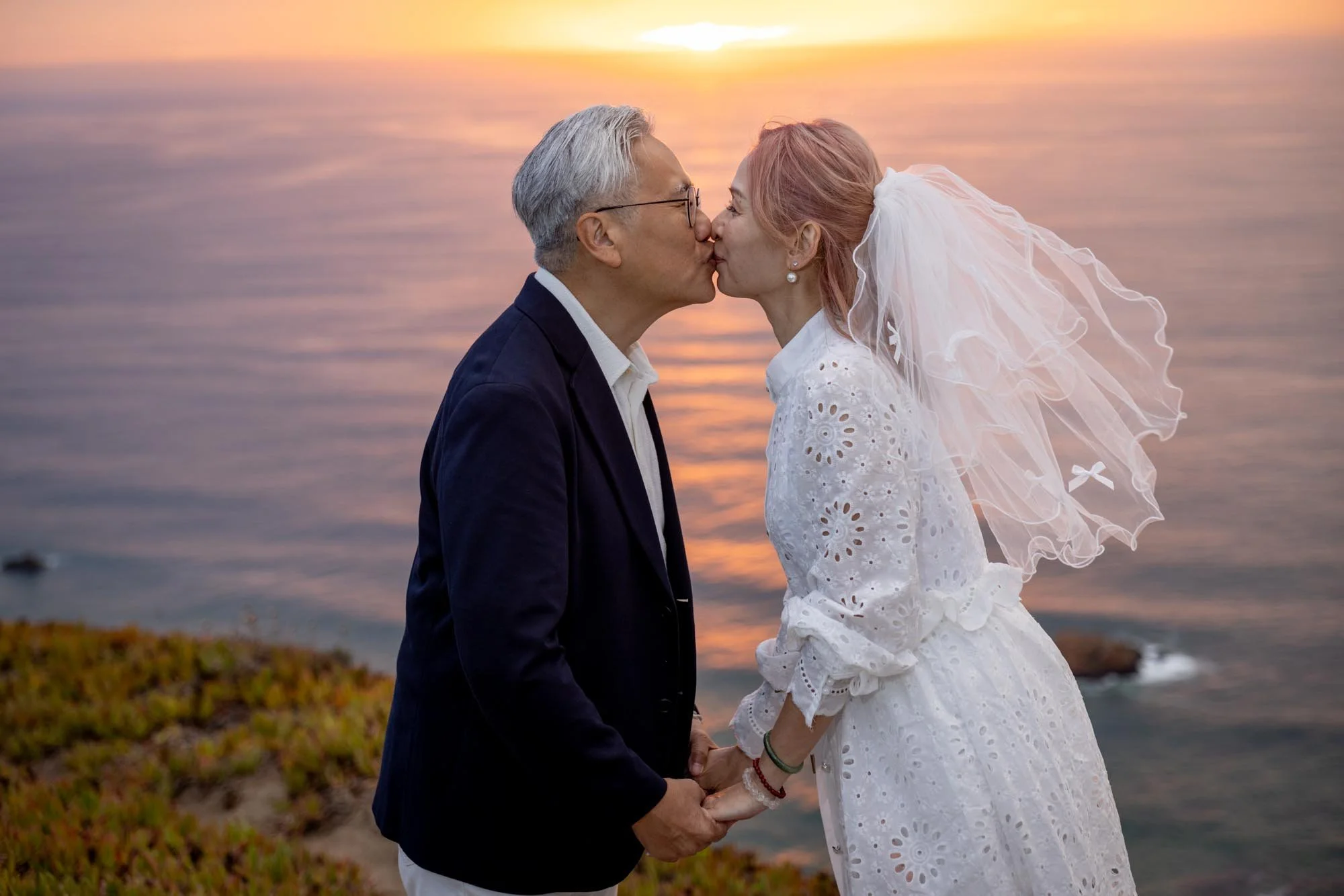A couple, dressed in wedding attire, holding hands and kissing by the ocean during sunset.