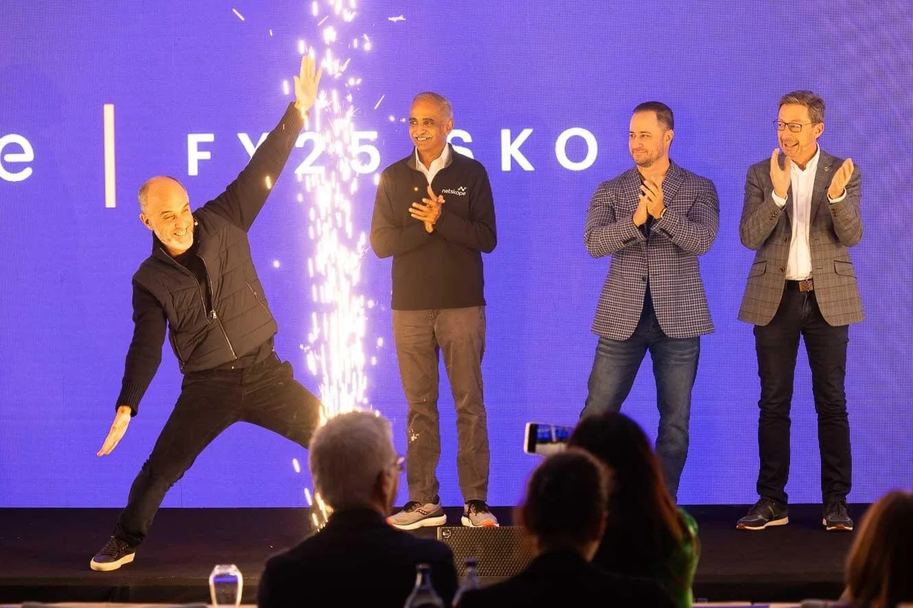 Four men on stage celebrating a successful event. One man is performing a dramatic pose with sparklers, while three others are clapping and smiling. The background displays the text 'FY 25 K O' on a blue screen.