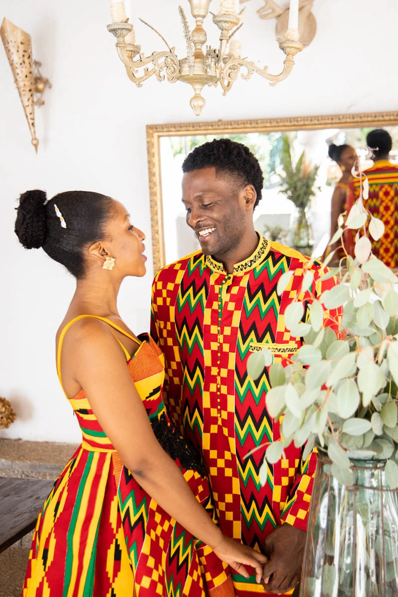 A couple dressed in colorful traditional African attire, holding hands and looking at each other affectionately in a warmly decorated room with a chandelier and a large mirror.