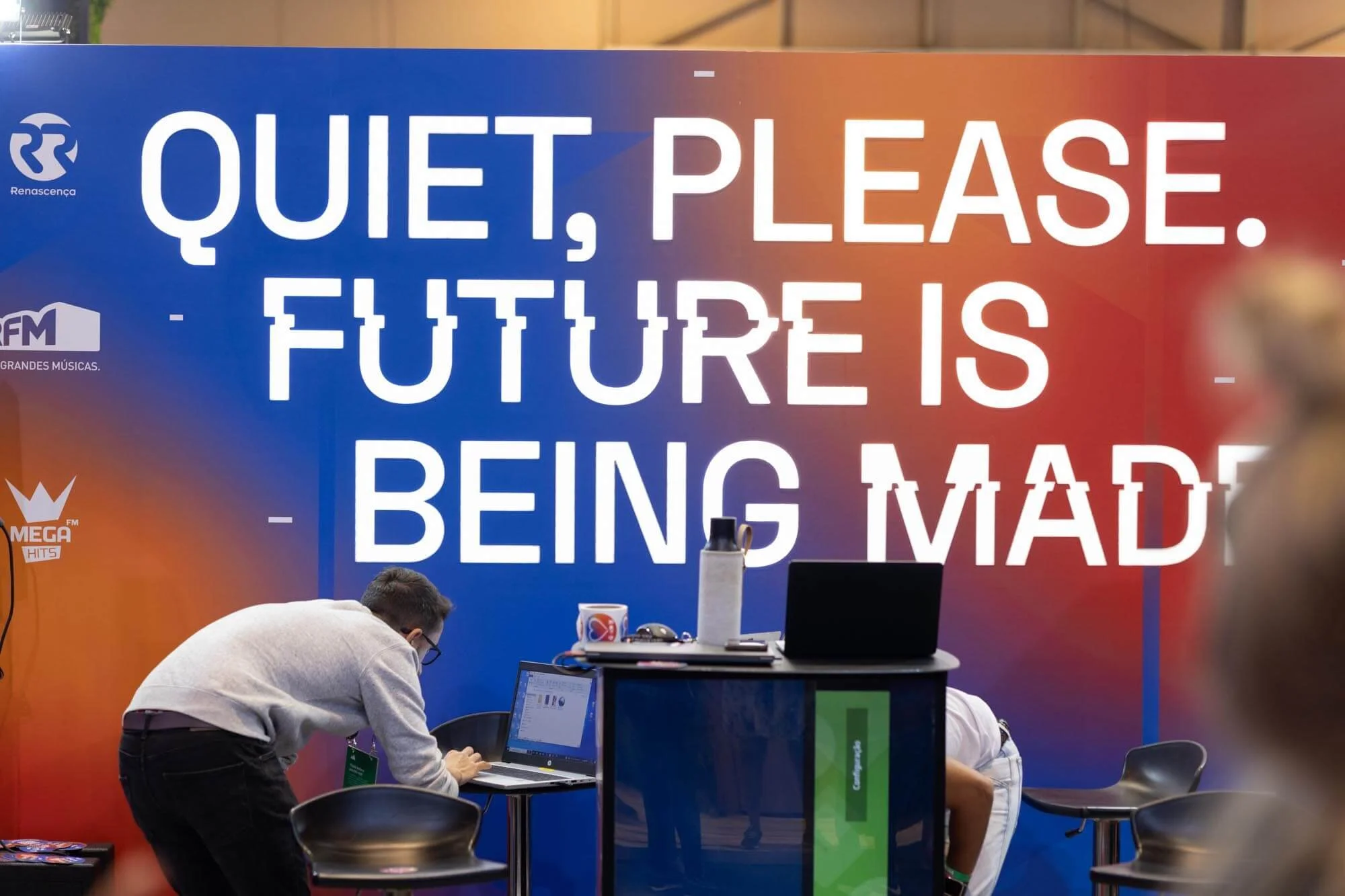 A large sign with blue and red background and white text that reads, "Quiet, please. Future is being made." In front of the sign, a person in glasses and a light gray sweater is working on a laptop at a small table, with a water bottle, mug, and othe