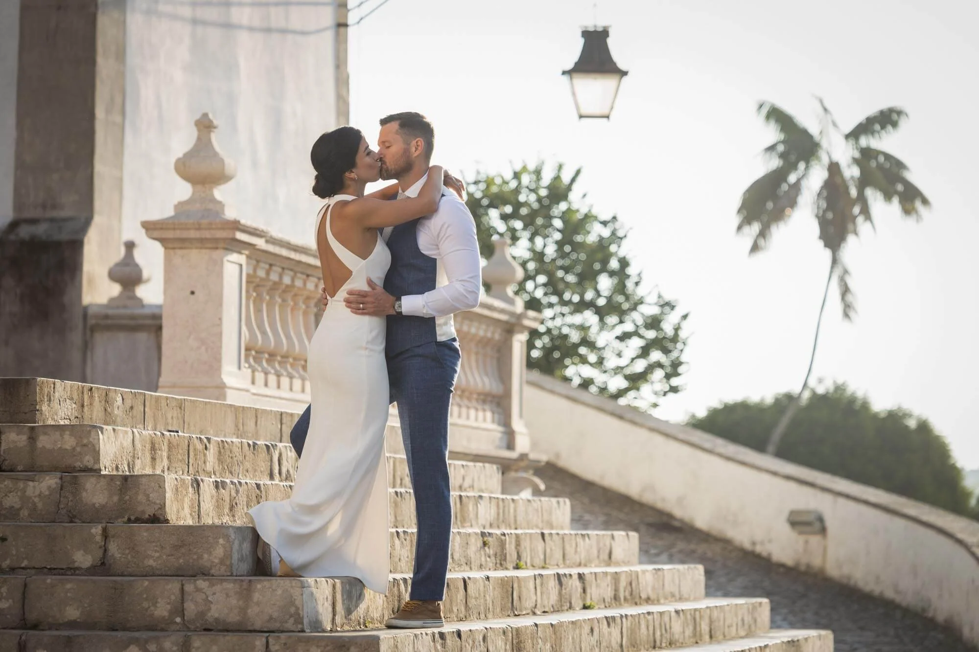A couple in wedding attire sharing a kiss on outdoor stone steps with a balustrade, a lamp post, and palm trees in the background, at Sintra national palace.