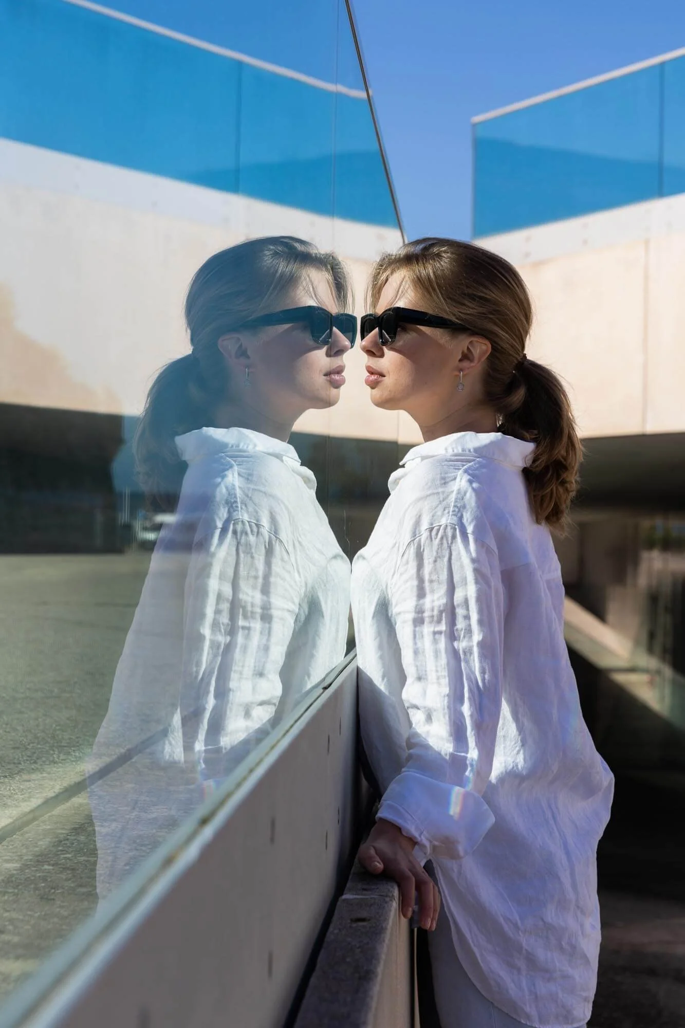 A woman in a white shirt and sunglasses is standing next to a glass wall, with her reflection visible in the glass. The scene is outdoors with a blue sky and modern building behind her.