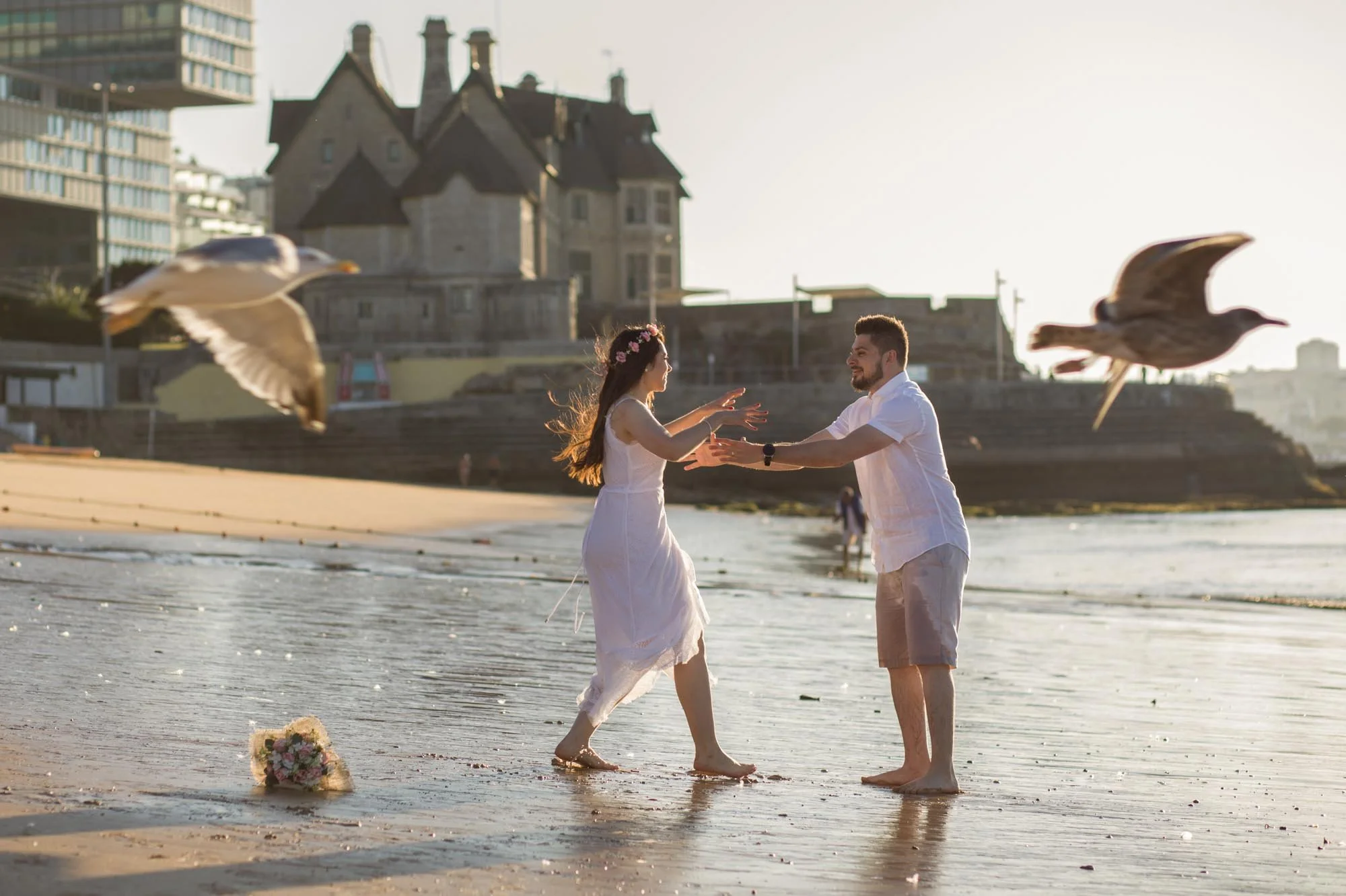 A couple on the beach during sunset, holding hands and smiling, with seagulls flying nearby and a bouquet of flowers on the sand.
