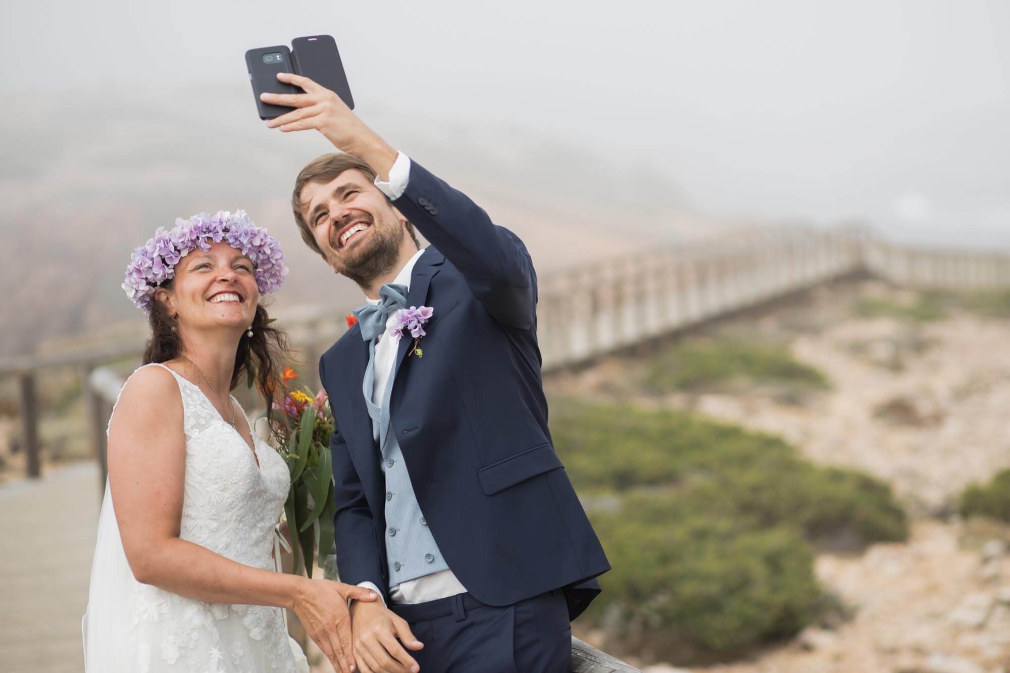 A happy bride and groom taking a selfie outdoors on a cloudy day, with a cloudy sky and a bridge in the background. The bride is wearing a white lace dress and a purple flower crown, holding a bouquet of flowers. The groom is wearing a dark blue suit