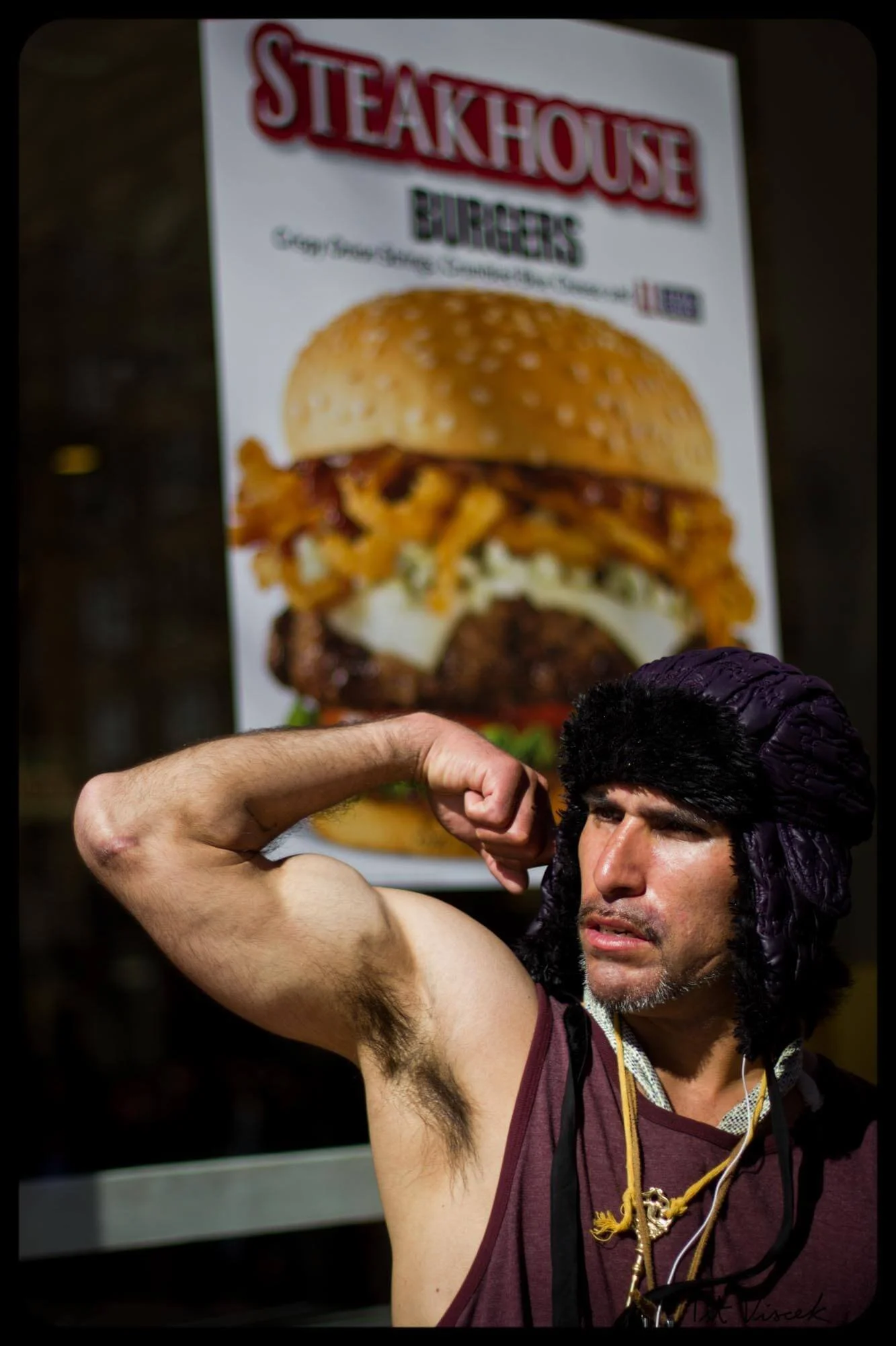 A man with dark hair and a beard wearing a dark fur hat and a sleeveless shirt flexes his bicep in front of a large poster advertising a burger. The poster features an image of a burger with fried chicken, cheese, and other ingredients.