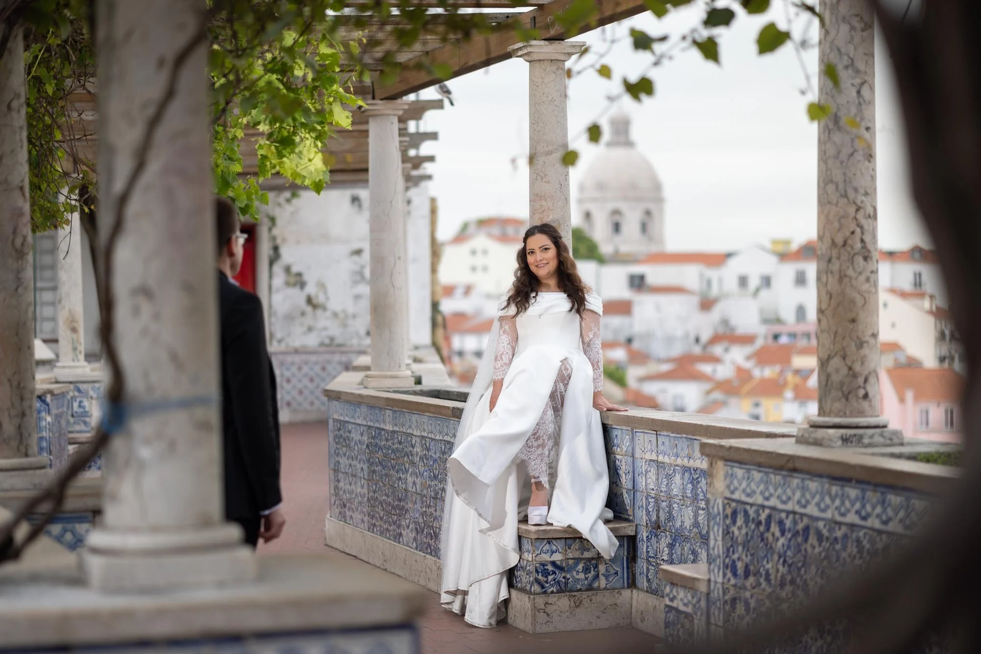 A woman in a white dress and lace sleeves sitting on a tiled ledge, smiling, with a man in a dark suit partially visible in the foreground, overlooking a cityscape with red-roofed buildings and a domed church in the background.