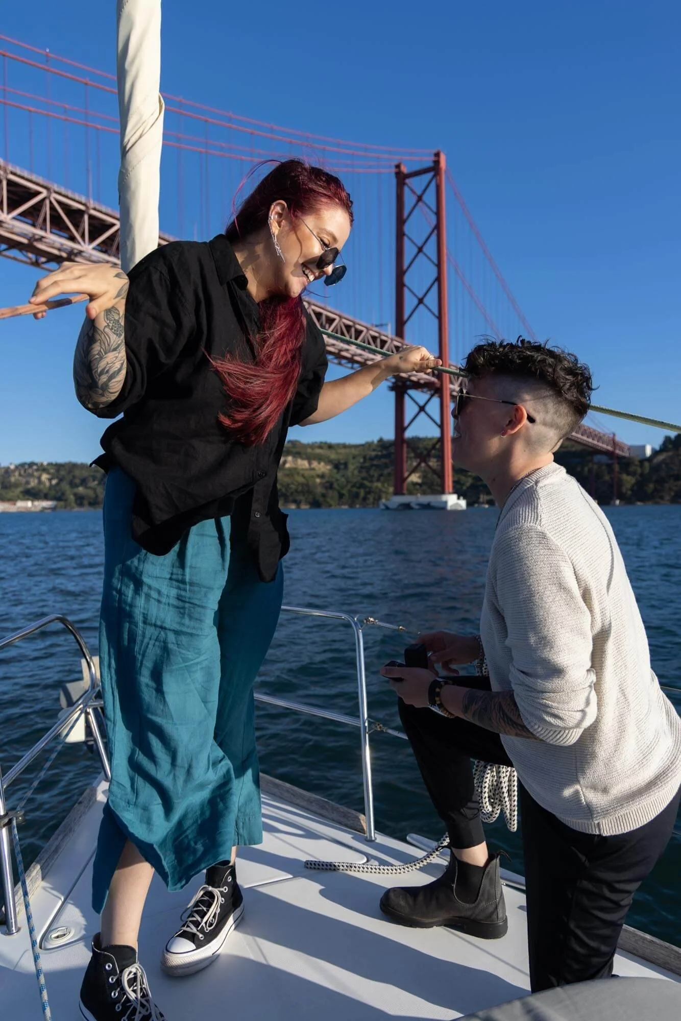A woman with tattoos and red hair smiling and leaning over the side of a boat, holding a rope, while a man in sunglasses kneels with a device in hand, near the Golden Gate Bridge on a sunny day.