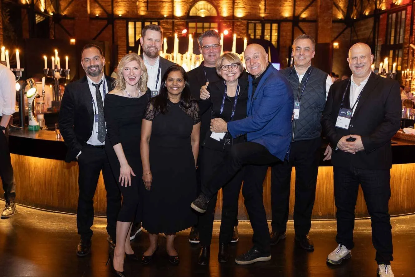 Group of nine people in formal attire, smiling and posing together at an indoor event with warm lighting and candles in the background.