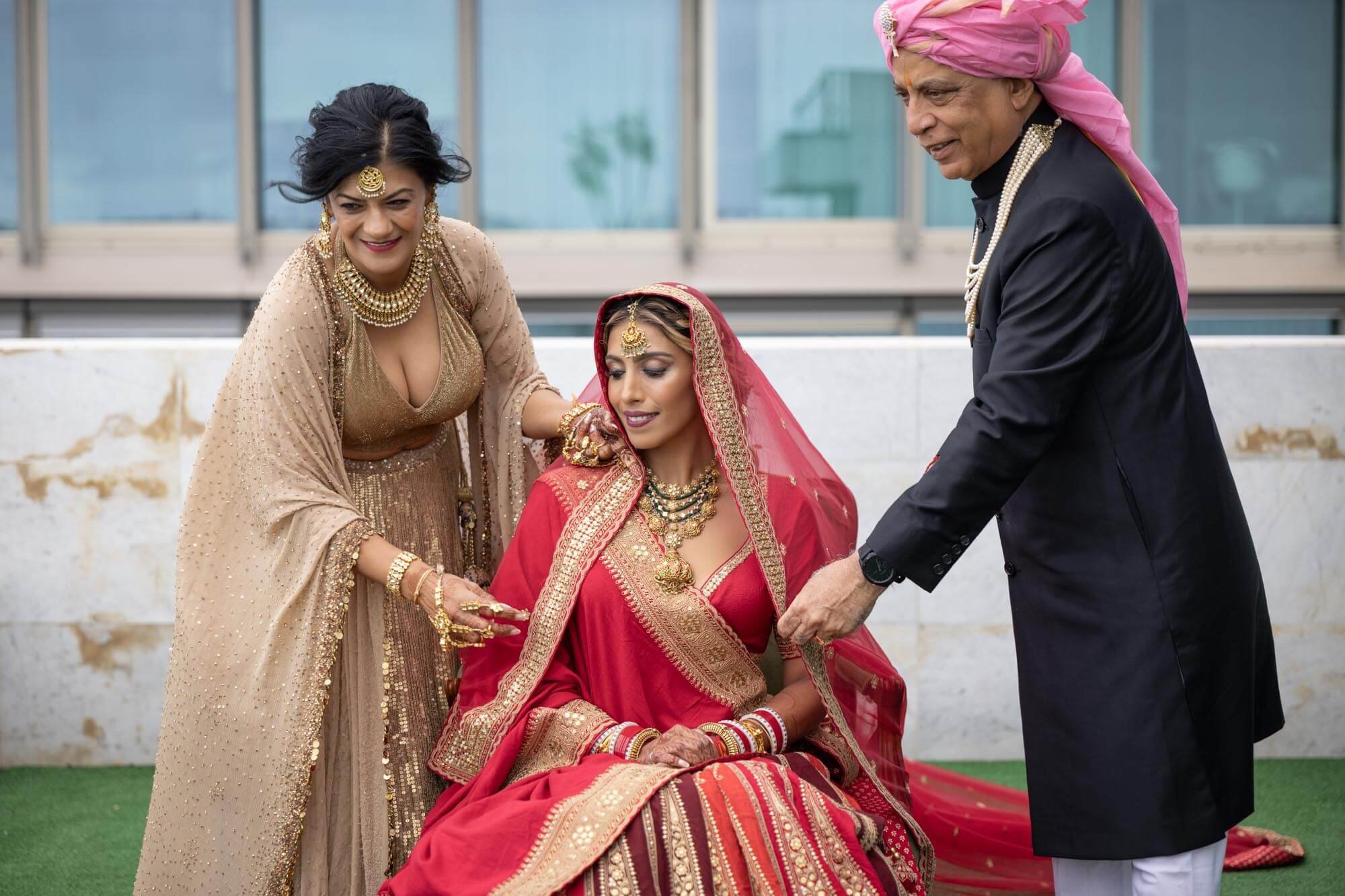 A woman dressed in red traditional Indian saree with jewelry, sitting on the ground with a gold necklace, bangles, and a headpiece, during a cultural or wedding ceremony. Two older people, a woman in gold attire and a man in black with a pink head co