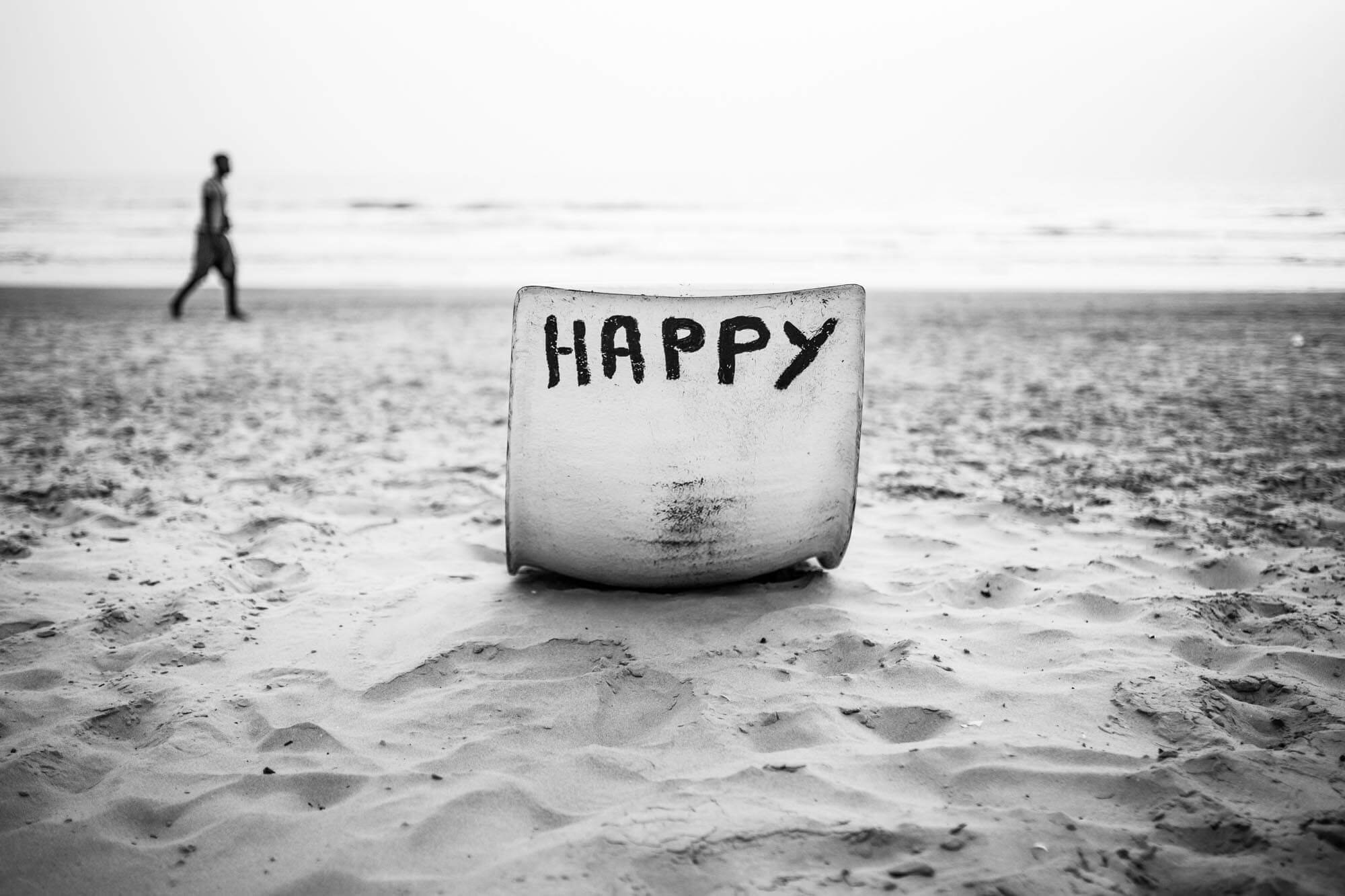A snow or ice sculpture with the word "HAPPY" on it, placed on the sand of a beach, with a person walking along the shoreline in the background in black and white.