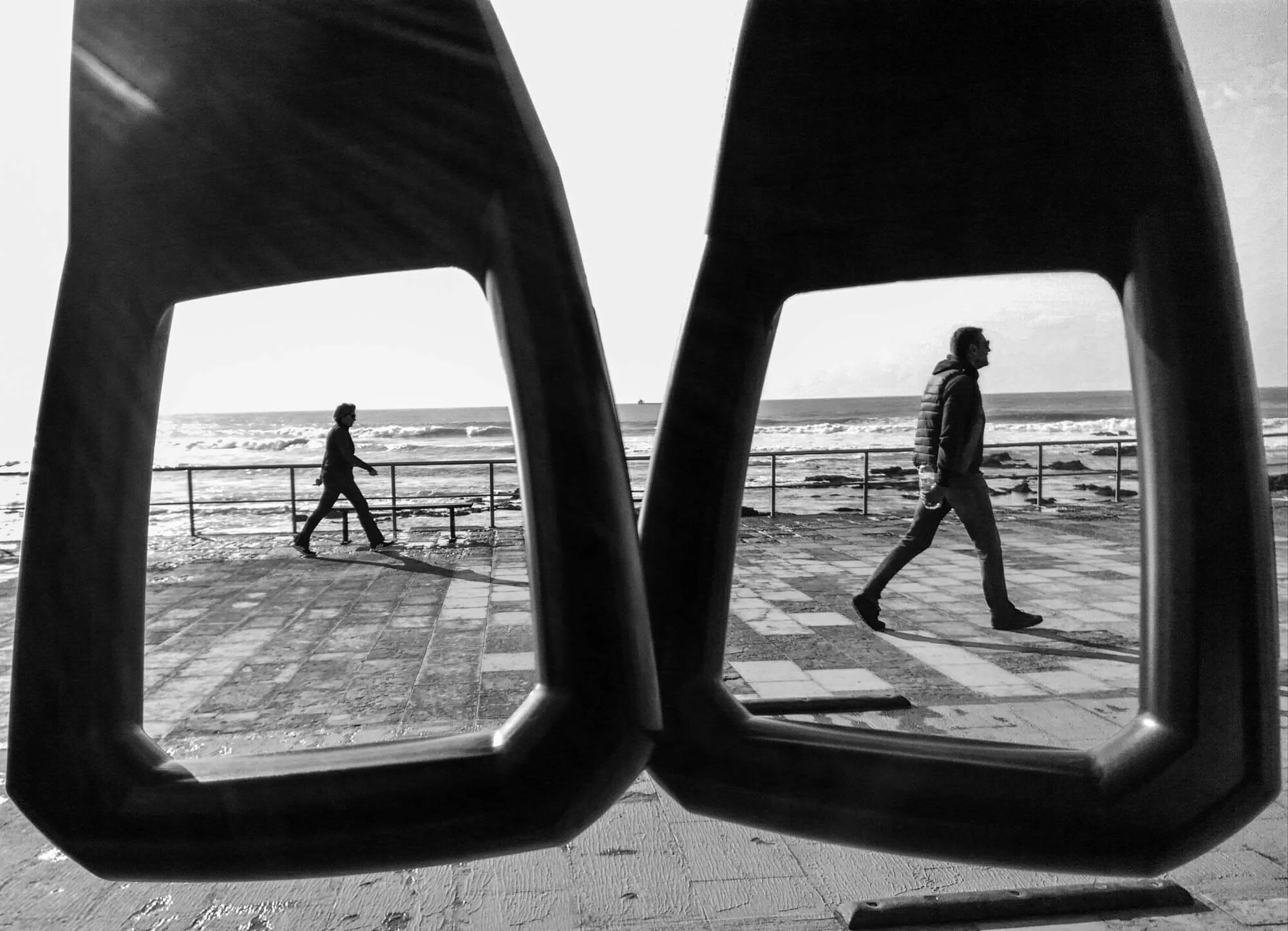Black and white photo of two people walking along a seaside promenade, seen through the lenses of a pair of glasses.