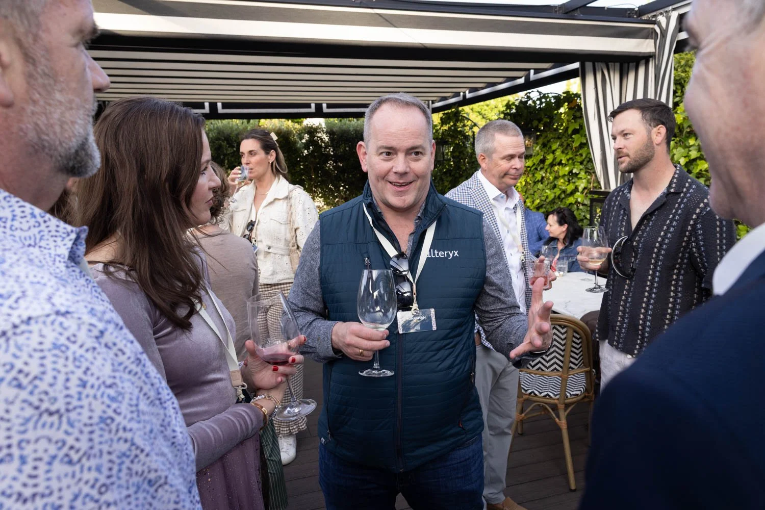 A group of people at a social gathering outdoors, with a man in the center wearing a blue vest speaking and holding a wine glass, others listening and holding wine glasses, with a canopy and greenery in the background. Posing for an Lisbon event phot