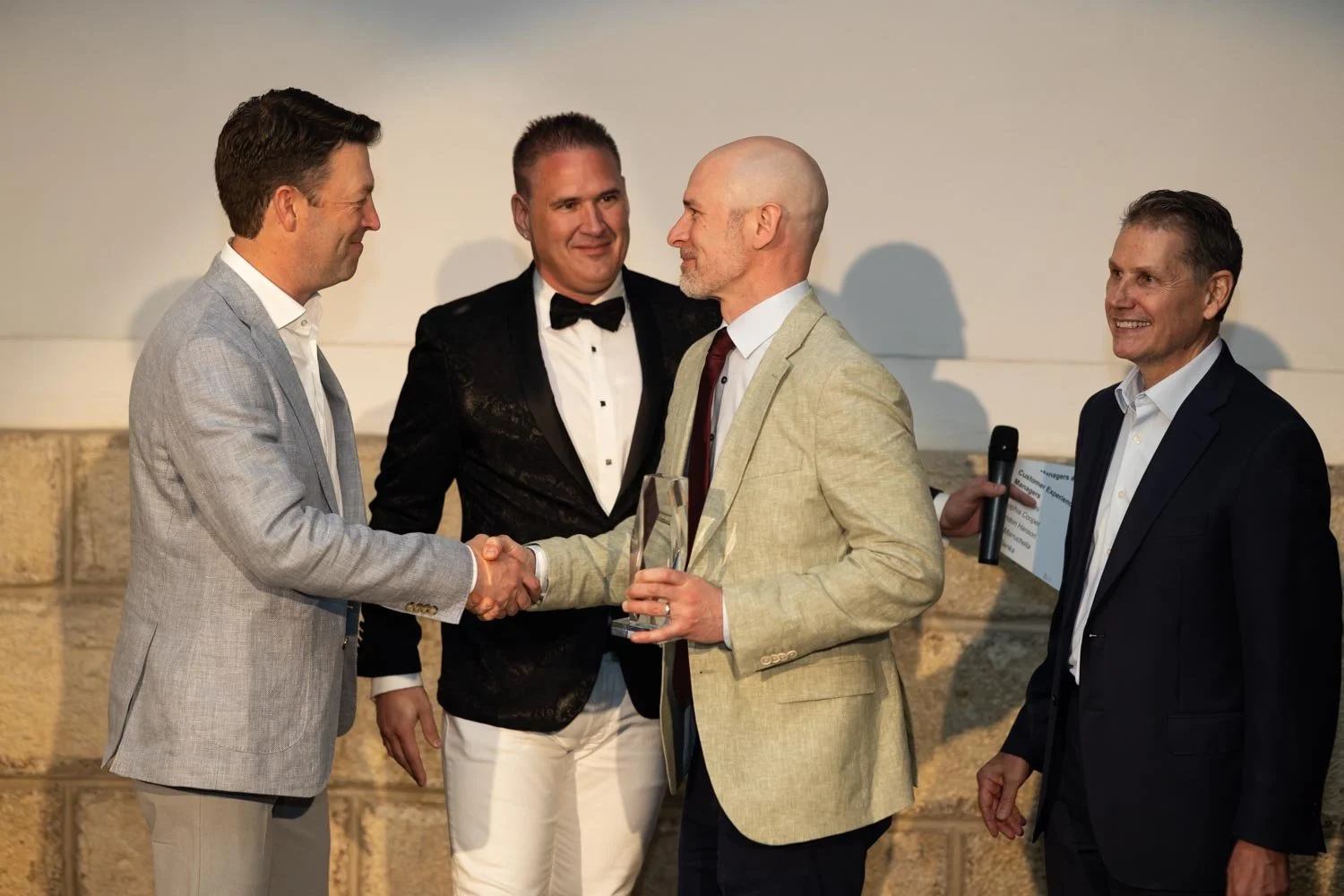 Four men in formal suits are at an award ceremony. Two of them are shaking hands, one is holding a glass award, and the fourth man is holding a microphone and a certificate. They are smiling and seem to be celebrating the award presentation.