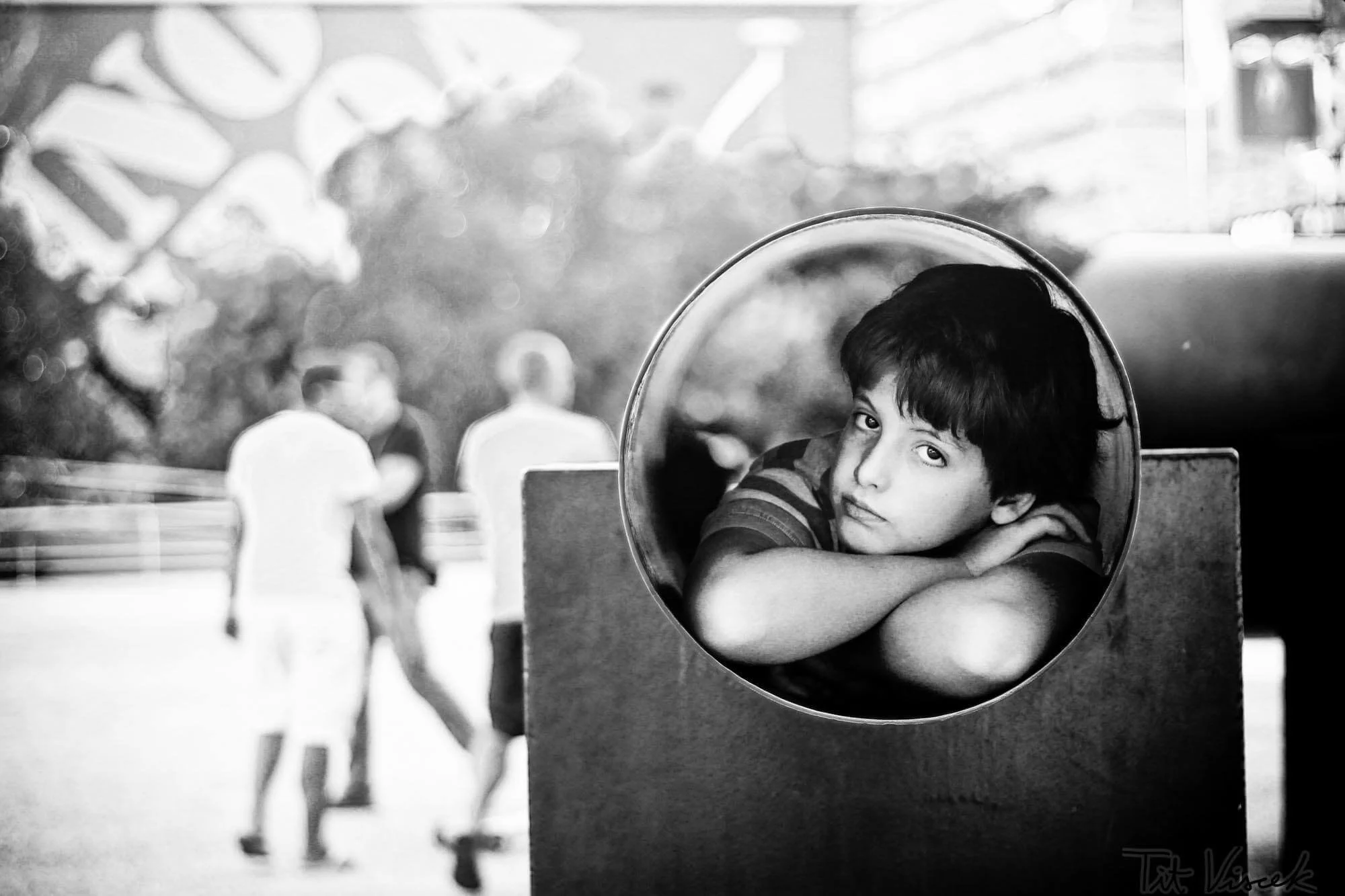 A boy resting his head on his arms, looking through a circular window at the camera. In the blurry background, three people are walking and talking. The image is in black and white.
