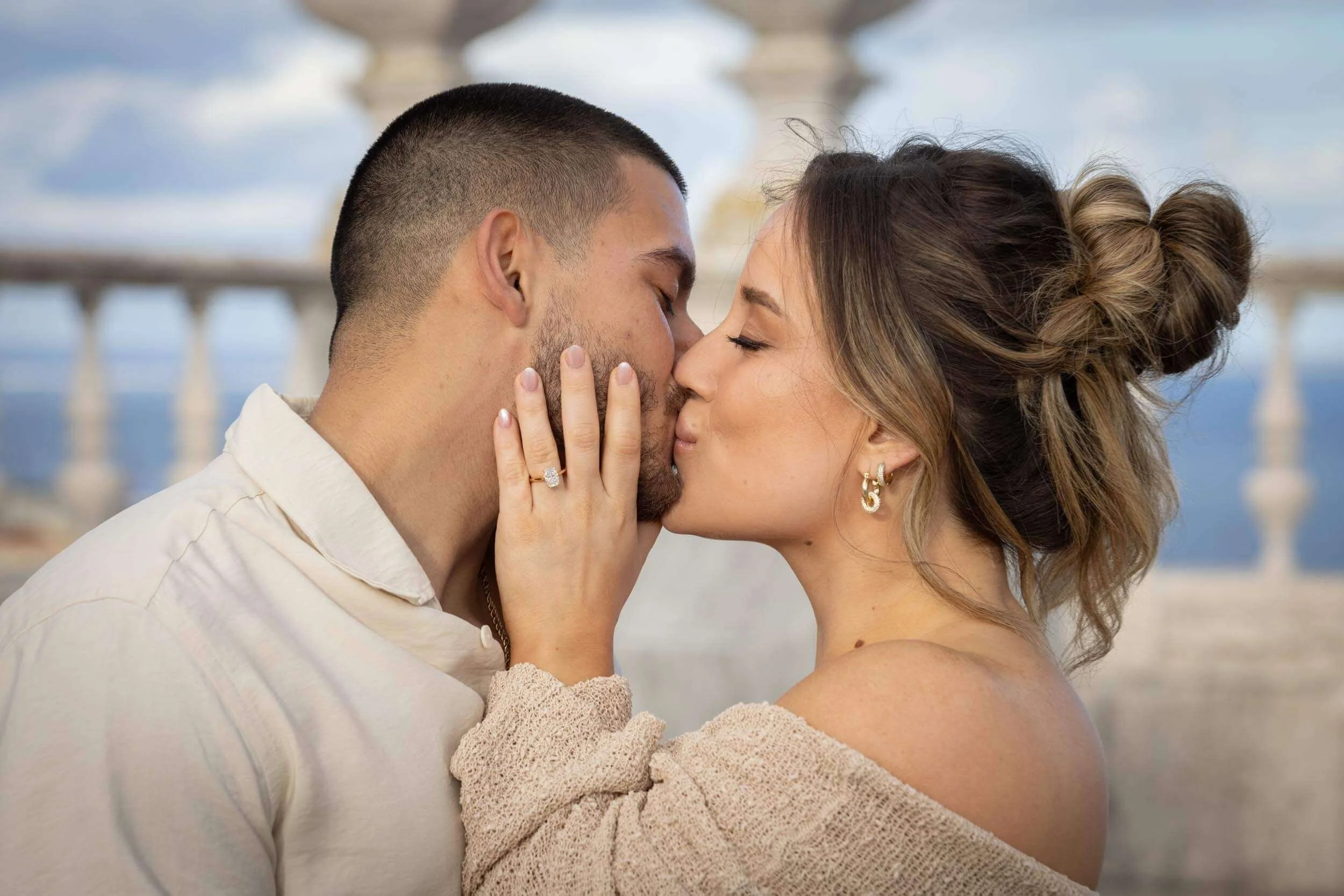 A man and woman kissing closely with their eyes closed, the woman's hand with an engagement ring touching the man's face, standing outdoors with a cloudy sky and architectural columns in the background.