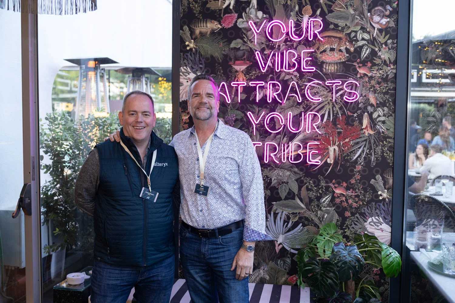 Two men standing indoors in front of a decorative wall with a neon sign that reads 'Your Vibe Attracts Your Tribe'. The man on the left is wearing a navy blue vest with a logo and the man on the right is wearing a white patterned shirt. They are smil