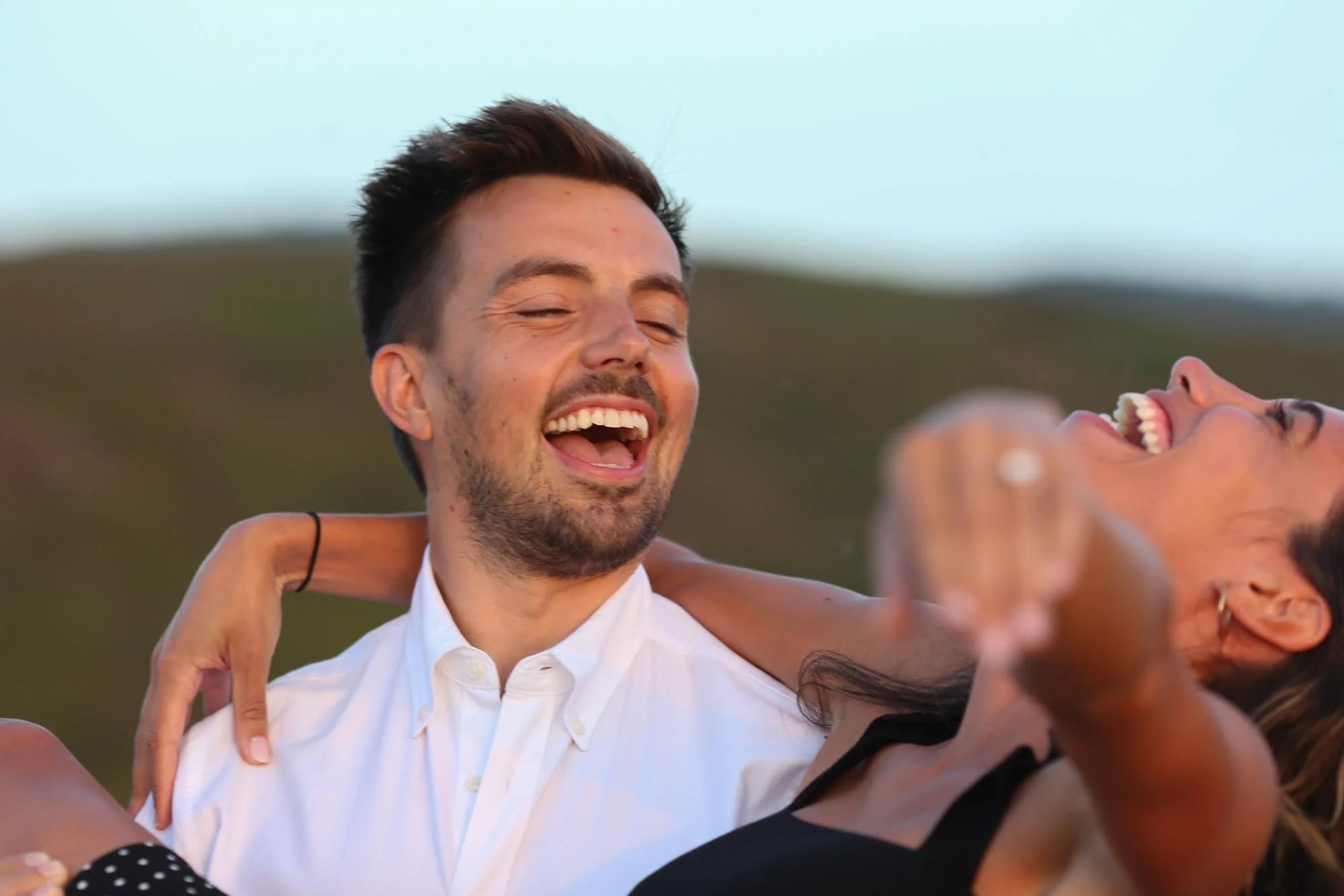 Two people are smiling and laughing, with one person's arm around the other's shoulders, outdoors during daytime.