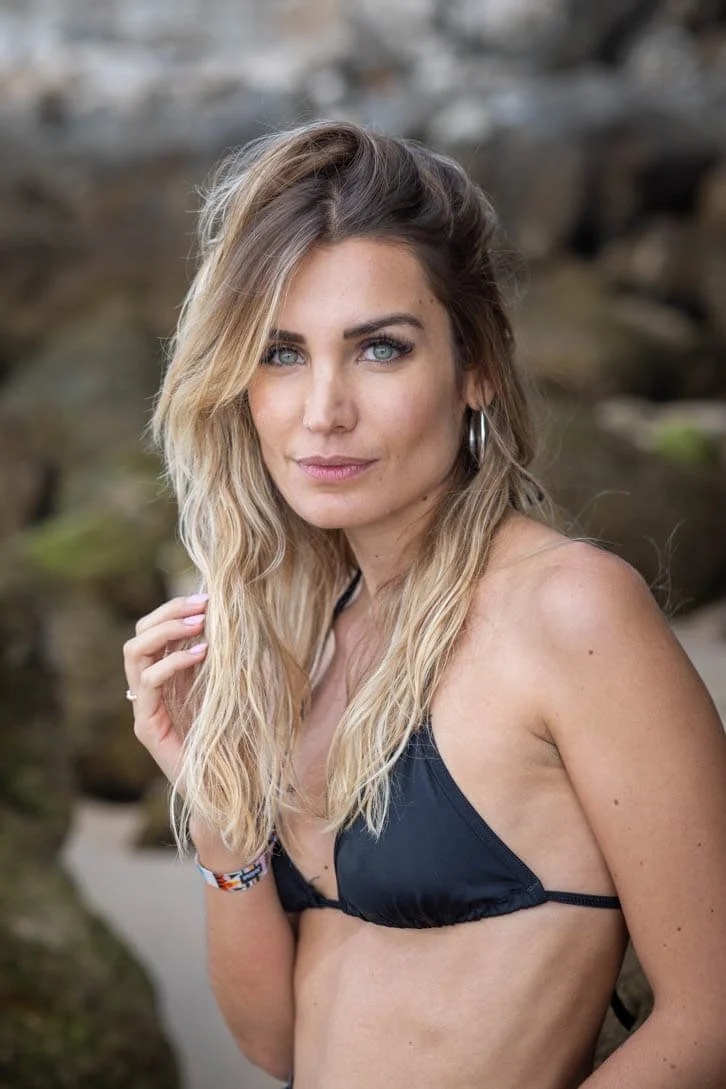 A woman with long, wavy blonde hair and blue eyes wearing a black bikini top on a beach with rocks in the background.