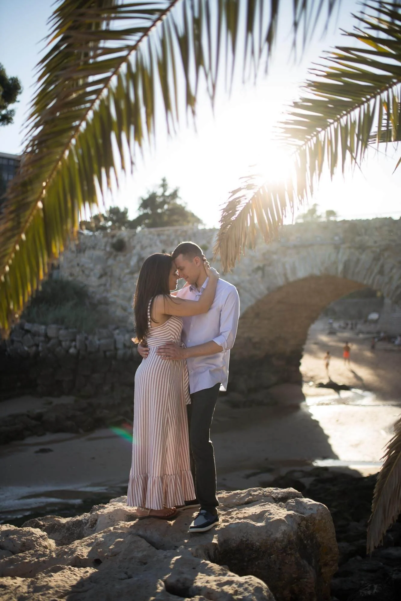 A couple embraces on a rocky beach with palm leaves in the foreground and an arch bridge in the background during sunset.