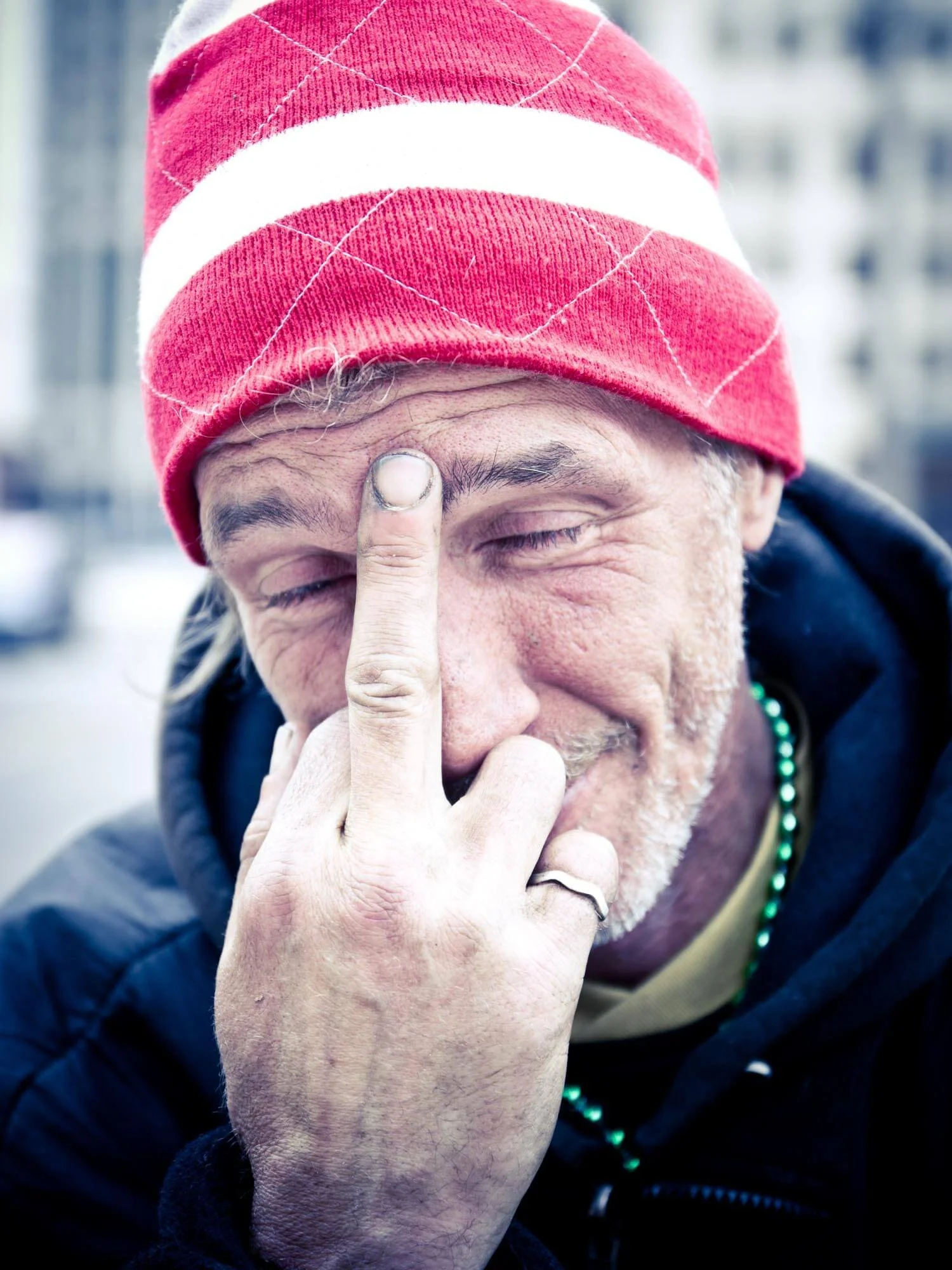 Man wearing red and white striped beanie, making a playful gesture with his hand near his face, outdoors with blurred buildings in the background.