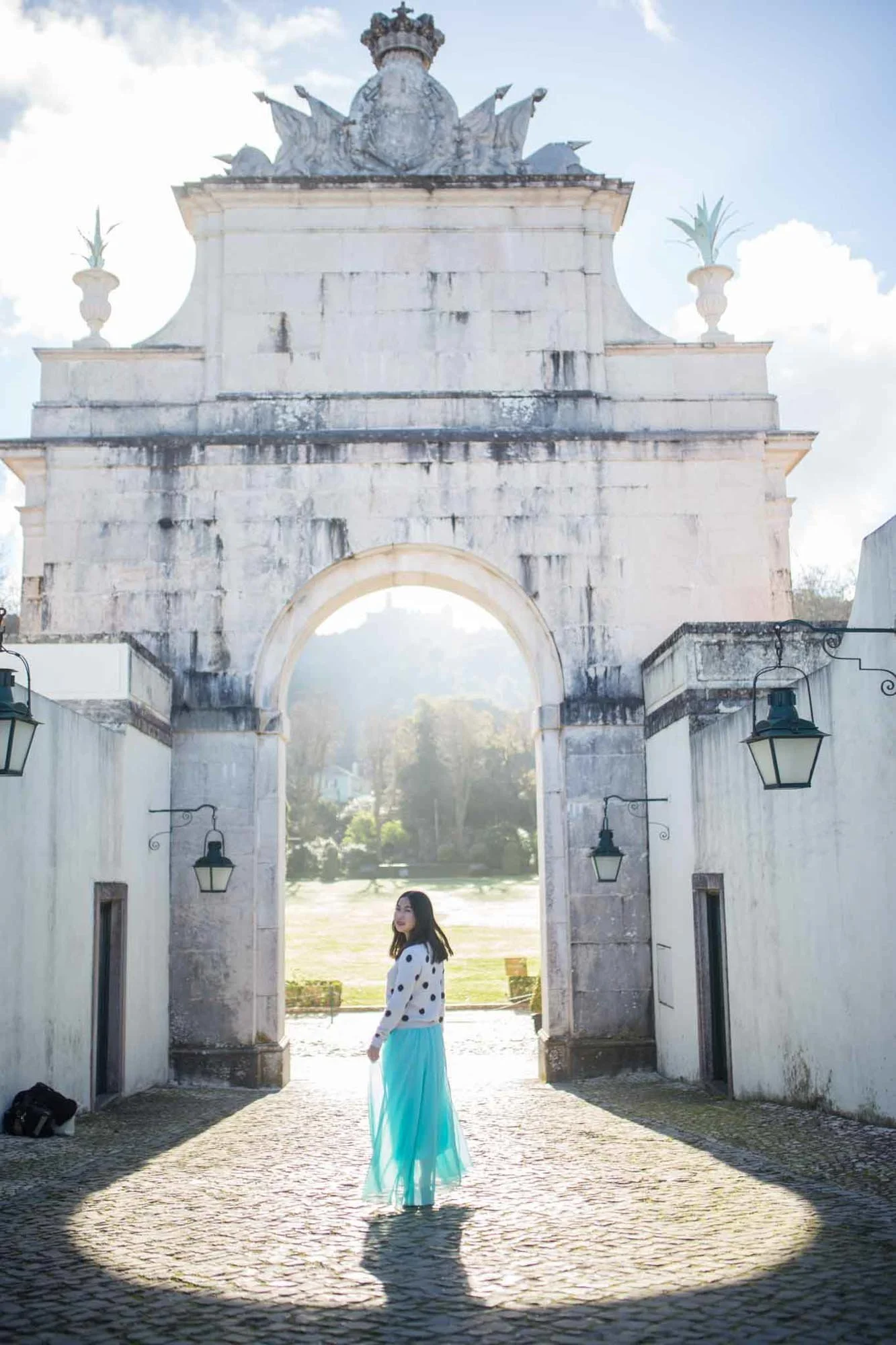 A woman standing in front of a large stone gate with an archway, and a tower-like structure on top, with a bright sunny sky in the background. She is wearing a black and white polka dot top and a long blue skirt.