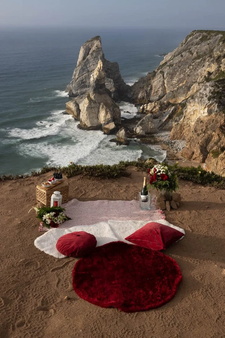 A romantic setup with pillows, rugs, flowers, and champagne on a sandy cliffside overlooking the ocean, with rocky cliffs of Cabo da Roca and waves in the background.